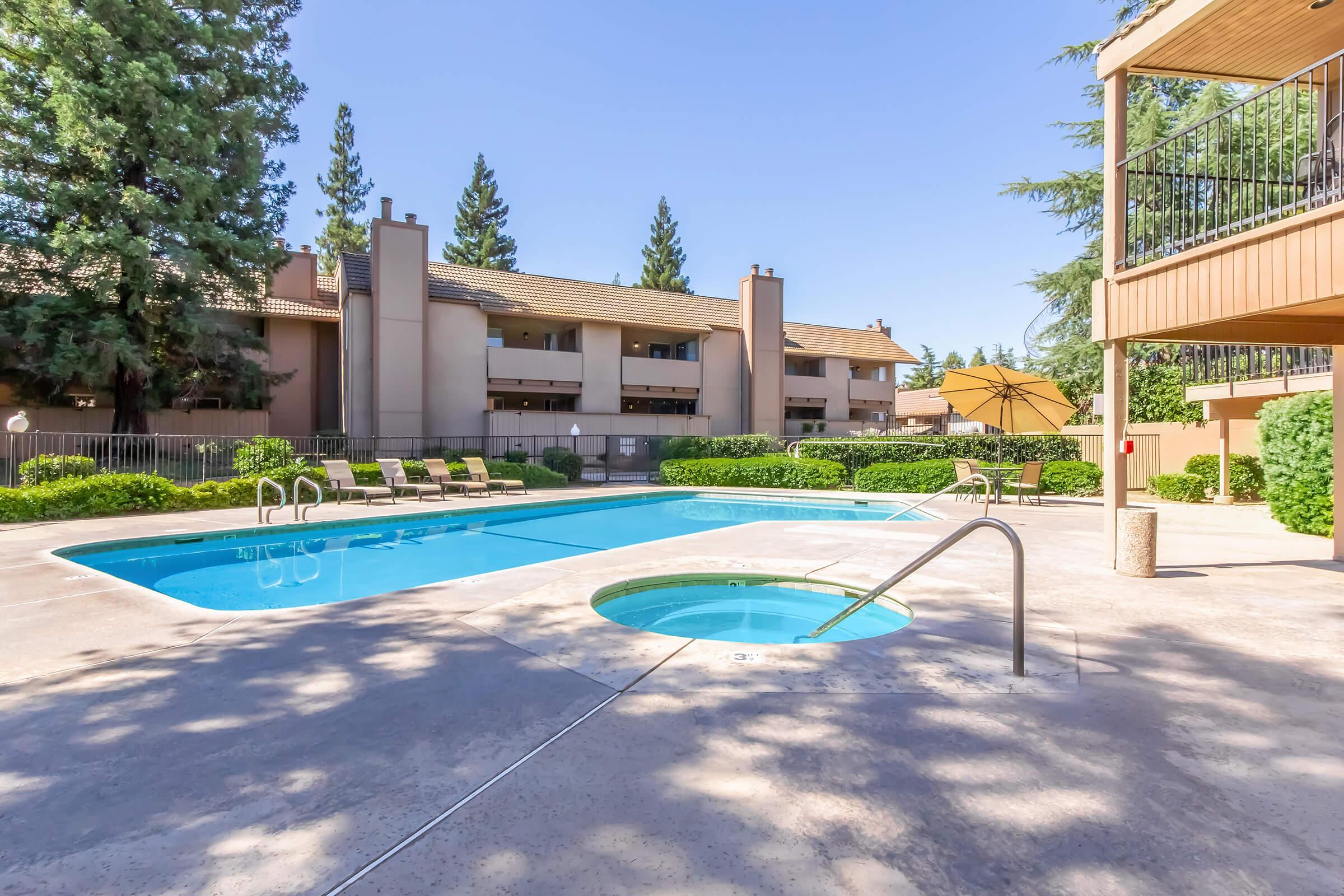 A serene outdoor pool area at an apartment complex, featuring a clear blue swimming pool, a hot tub nearby, and surrounded by lush greenery and trees. Several lounge chairs are placed around the pool, with a sunny sky above and buildings in the background.