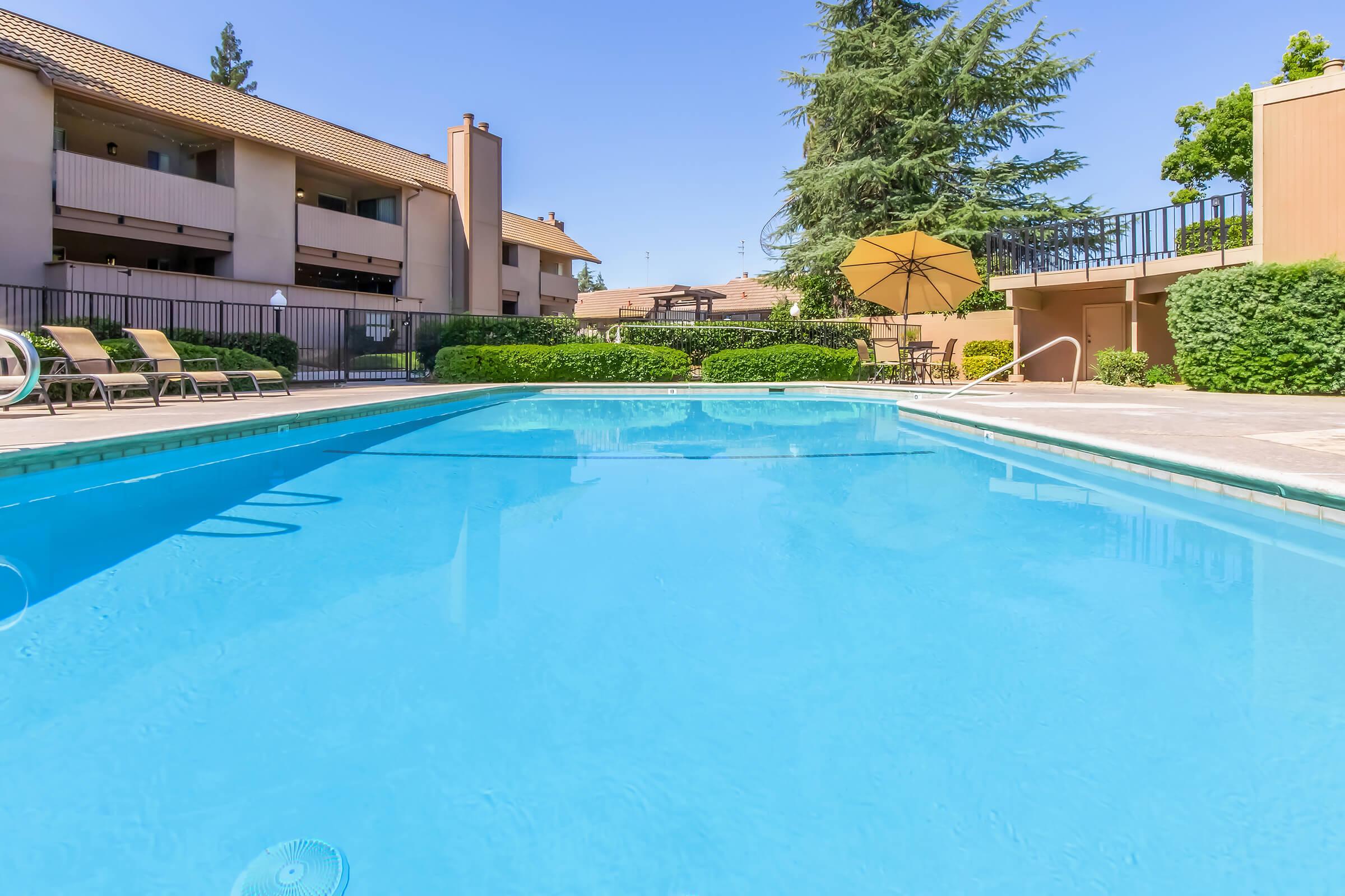 A clear blue swimming pool surrounded by lounge chairs and greenery, with a patio area featuring an umbrella. The sky is bright and sunny, and there are townhouses visible in the background.