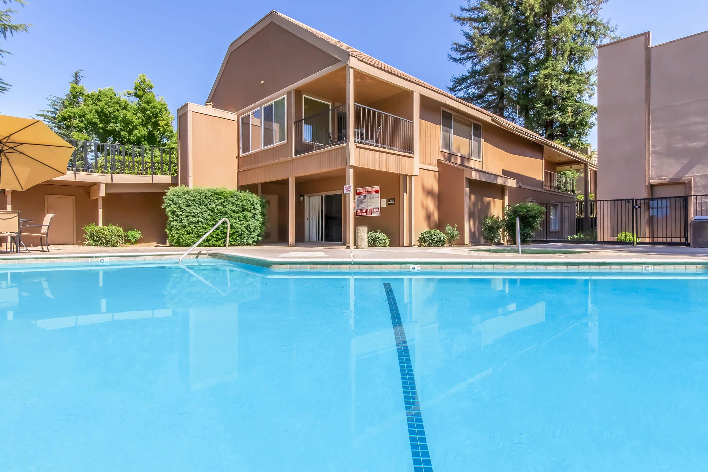 A clear swimming pool in the foreground with a two-story apartment building in the background. The building features balconies and is surrounded by green trees and shrubs. An umbrella and patio furniture are visible near the pool area, creating a relaxing outdoor atmosphere.