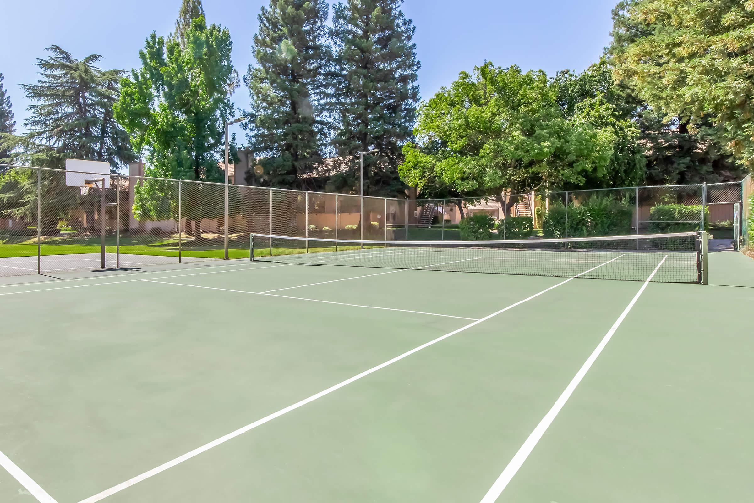 A well-maintained outdoor tennis court featuring green asphalt and a net, surrounded by tall trees and a grassy area. The court is empty, with clear blue skies overhead and a basketball hoop visible in the background, indicating a recreational area.