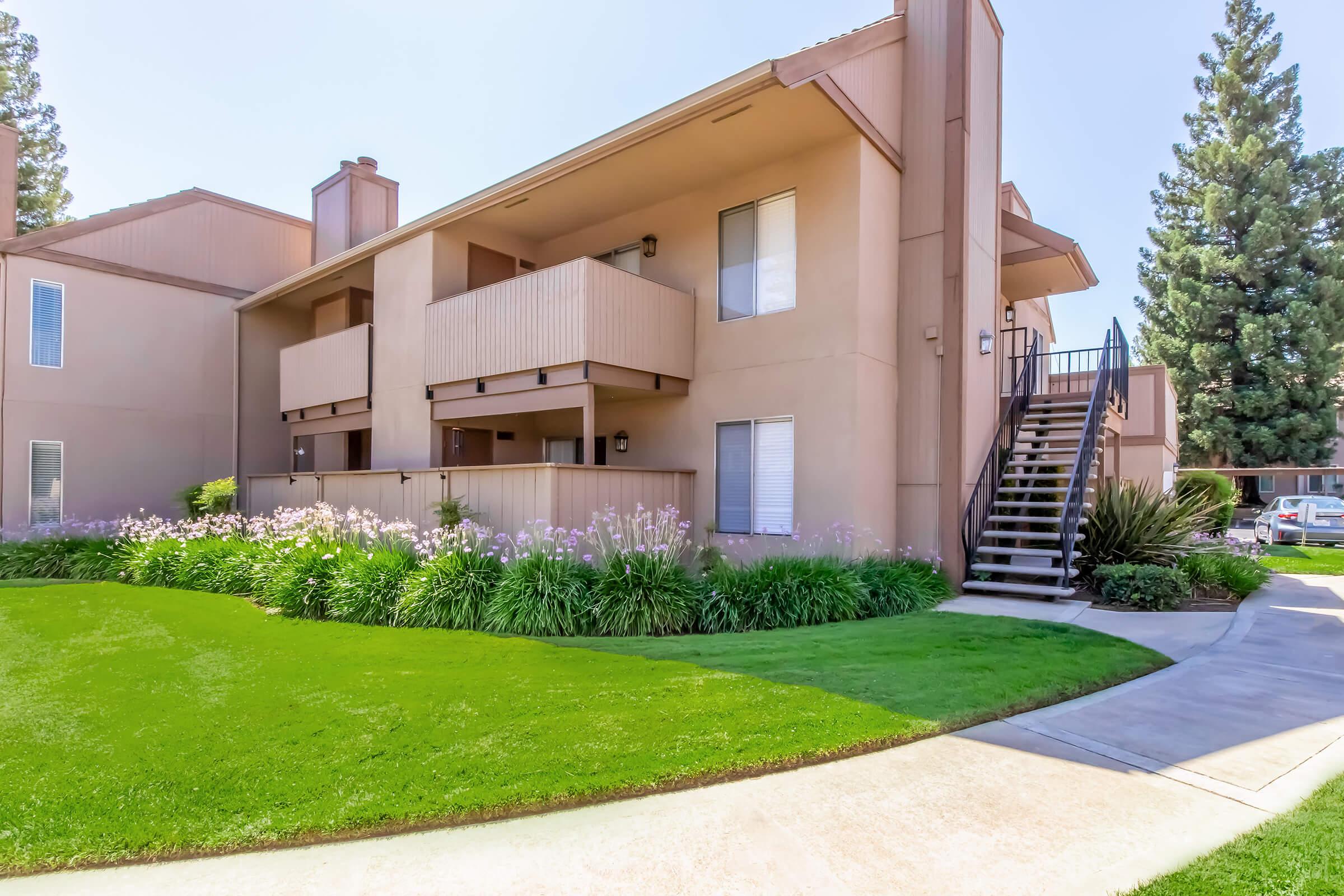 Two-story apartment building with a tan exterior and wooden balconies. Lush green lawn and flowering plants in front. A staircase leads to the upper level, with tall trees in the background. The scene is well-lit, suggesting a sunny day.
