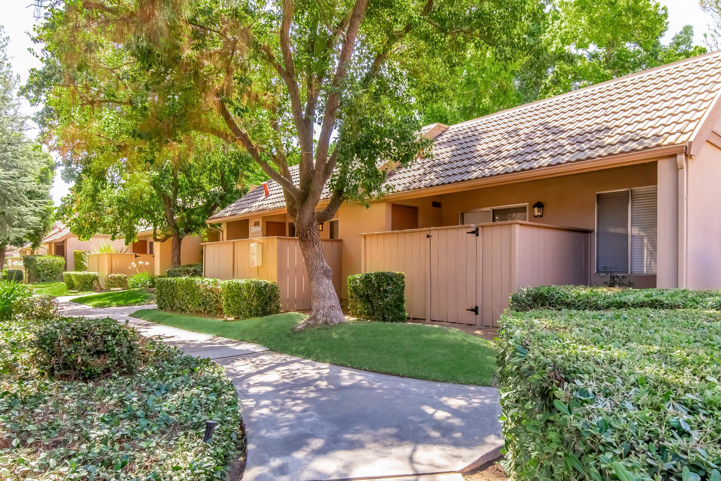 A pathway bordered by neatly trimmed hedges leads to several brown single-story buildings with tile roofs, nestled among lush green trees. The area appears well-maintained, with wooden fences enclosing outdoor spaces and ample natural light filtering through the foliage.