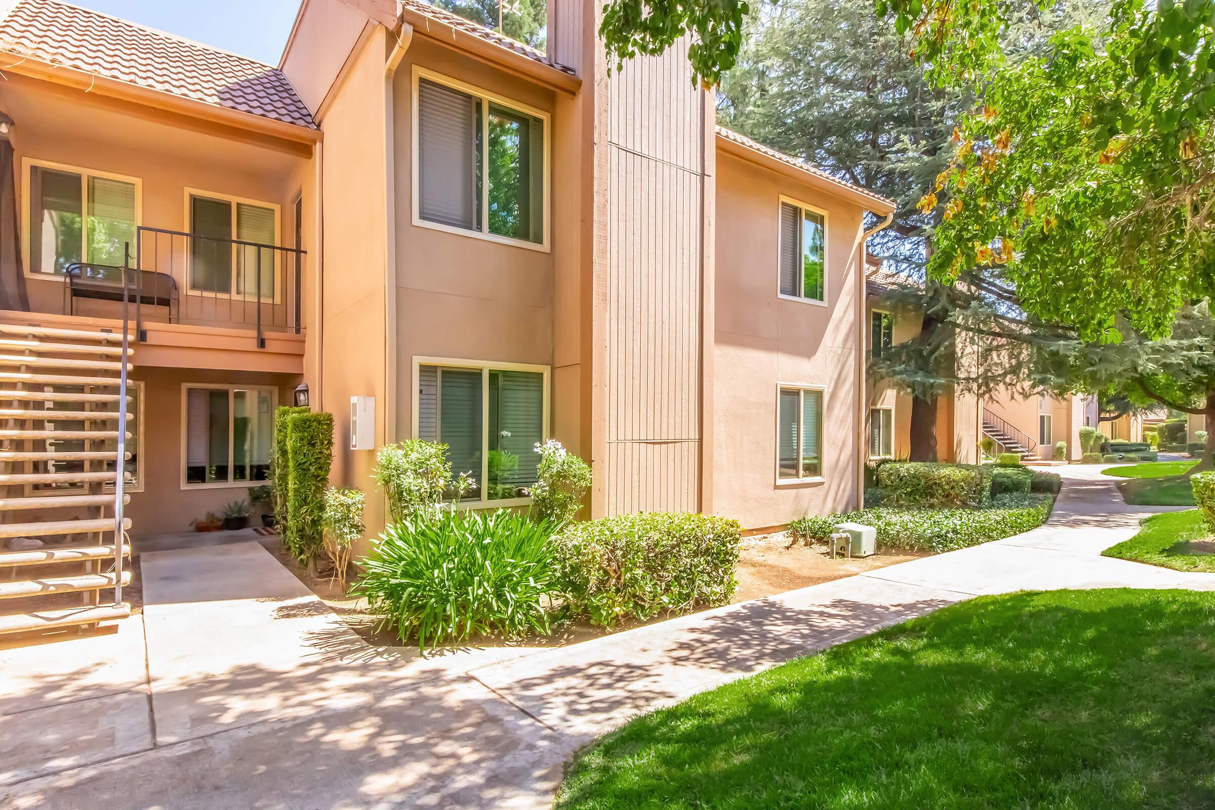 Exterior view of a multi-unit residential building featuring two levels, with a staircase leading to the upper floor. Surrounding the building are well-maintained greenery and a walkway, creating a pleasant residential environment. Bright sunlight enhances the overall appearance of the scene.