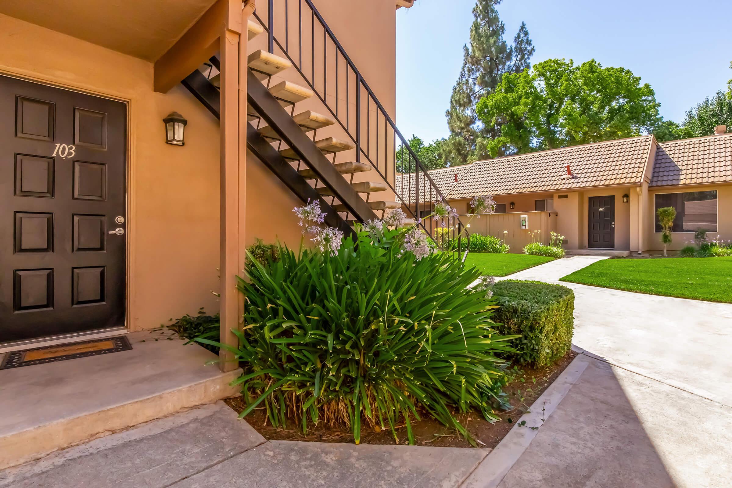 A well-maintained courtyard with green grass and flowering plants. A flight of stairs leads up to a door labeled "103." In the background, there are several other units with brown tiled roofs and lush trees, creating a serene and inviting atmosphere.