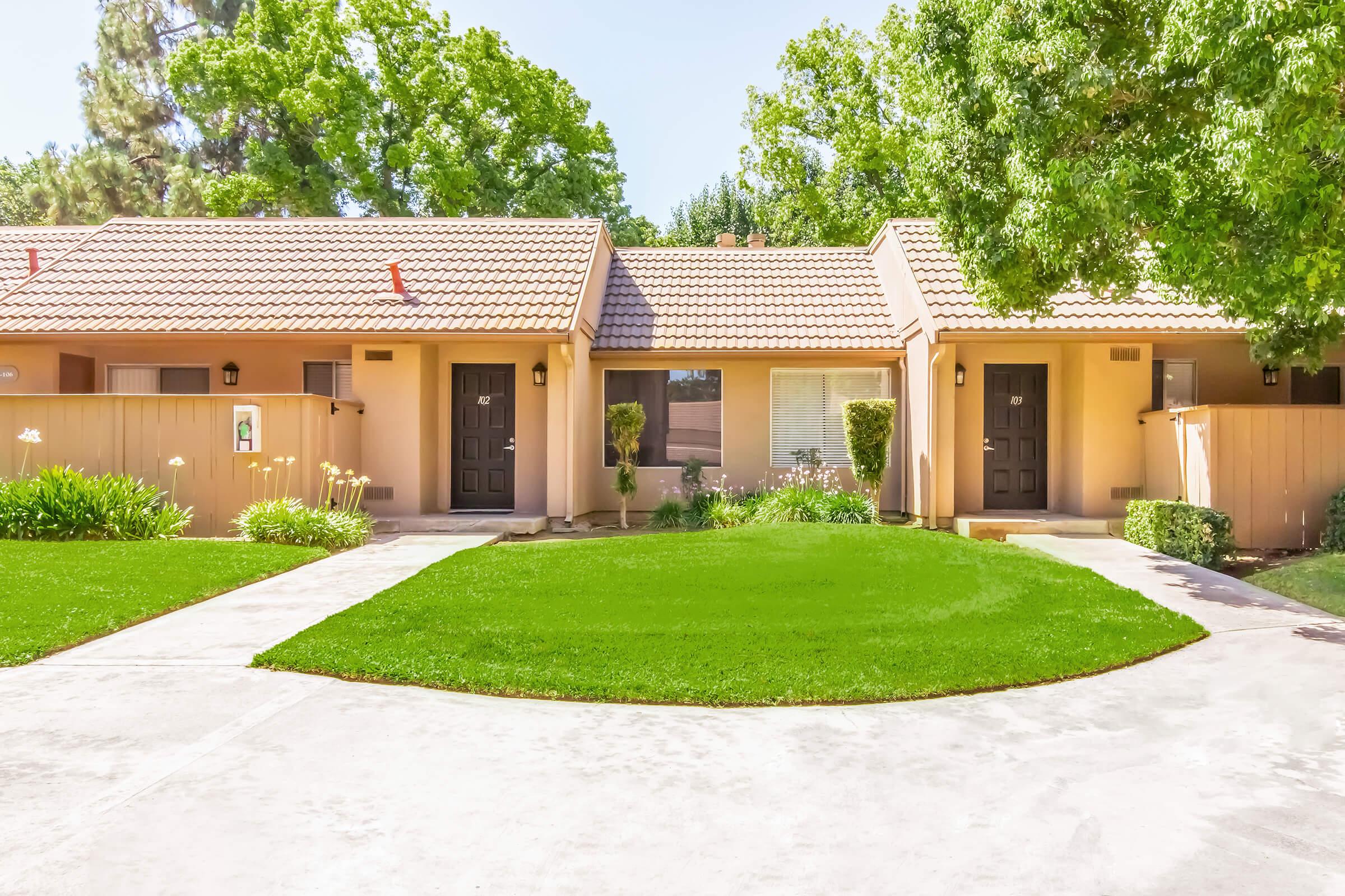 Two single-story residential units with brown doors, set in a well-manicured lawn. The units feature light-colored walls and terracotta tile roofs, surrounded by lush green bushes and trees. A clear pathway leads to each entrance, highlighting a tidy, inviting outdoor space.