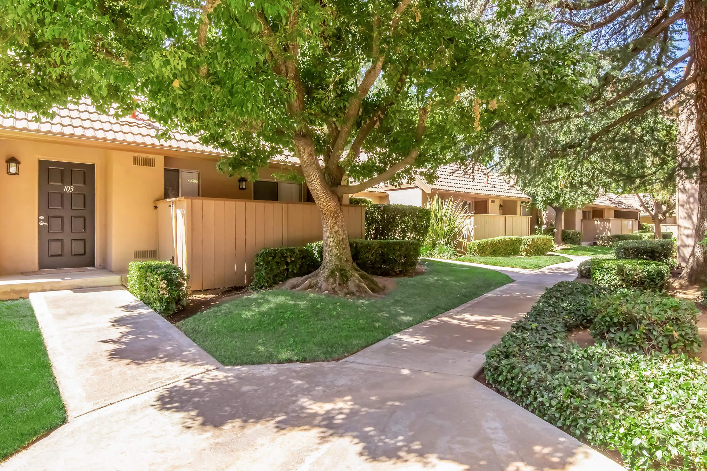 Walkway surrounded by well-manicured grass and hedges, leading to residential units with brown doors. A large tree provides shade, and the area is neatly landscaped, featuring pathways that connect the homes in a serene environment.