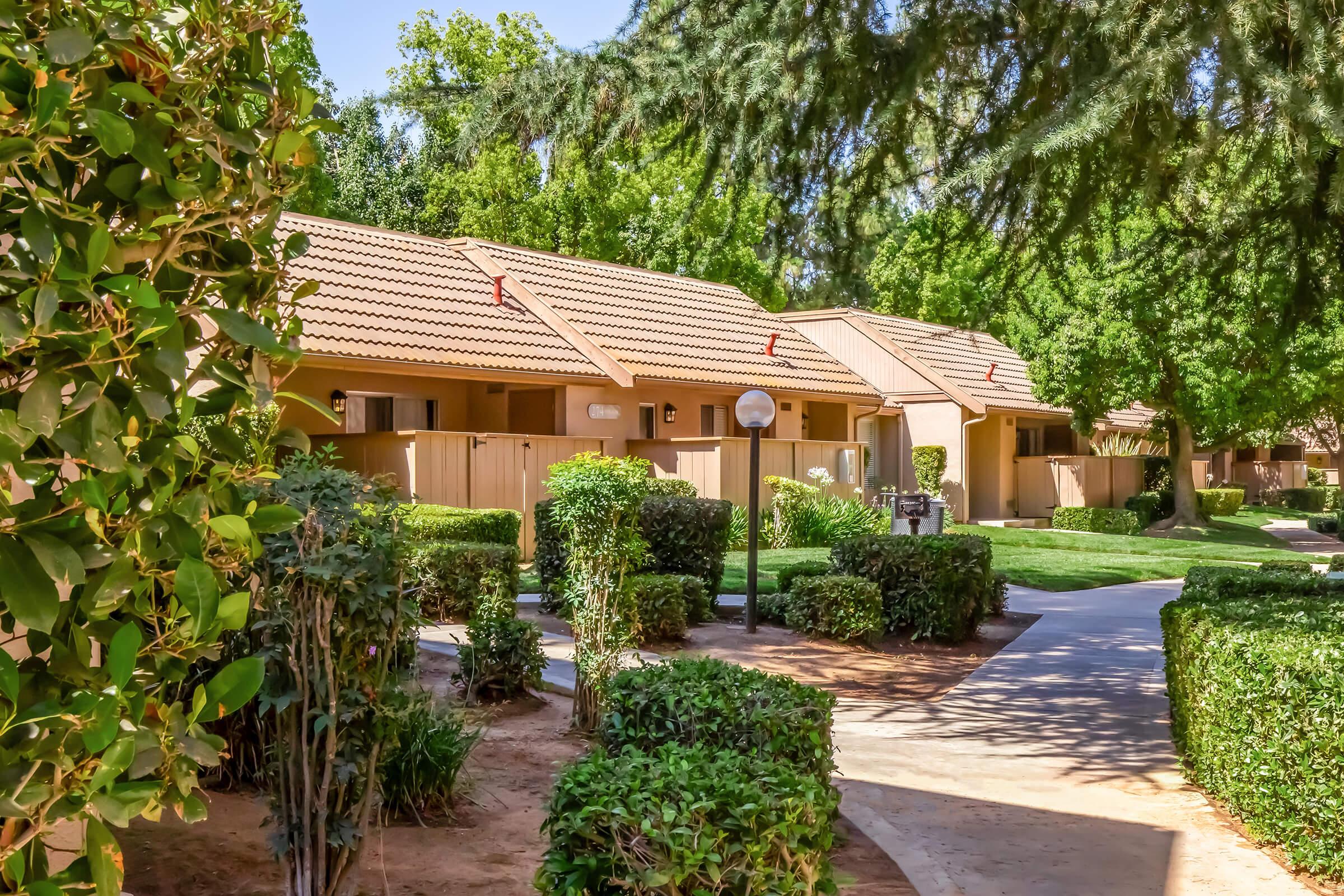 A peaceful landscaped area featuring several tan-colored buildings with tiled roofs. The scene includes well-maintained greenery, bushes, and pathways, creating a serene atmosphere. Soft sunlight filters through the trees, enhancing the inviting ambiance of the residential setting.