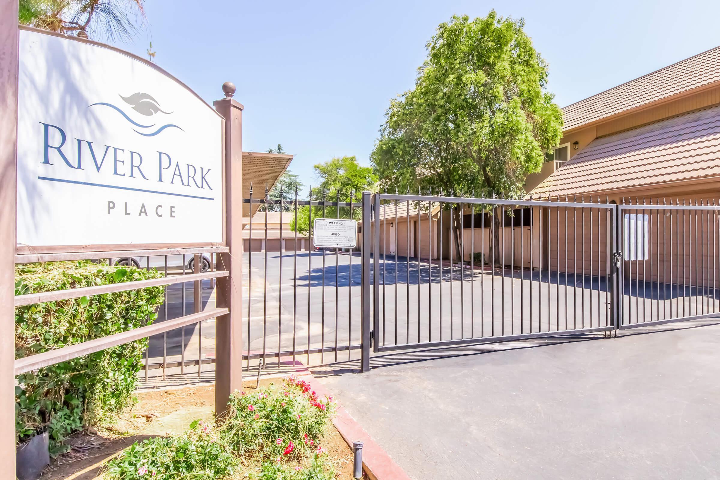 A gated entrance to River Park Place, featuring a sign with the name. The surrounding area includes greenery and a paved driveway leading into a parking space. The sky is clear, and the buildings in the background have a modern design.