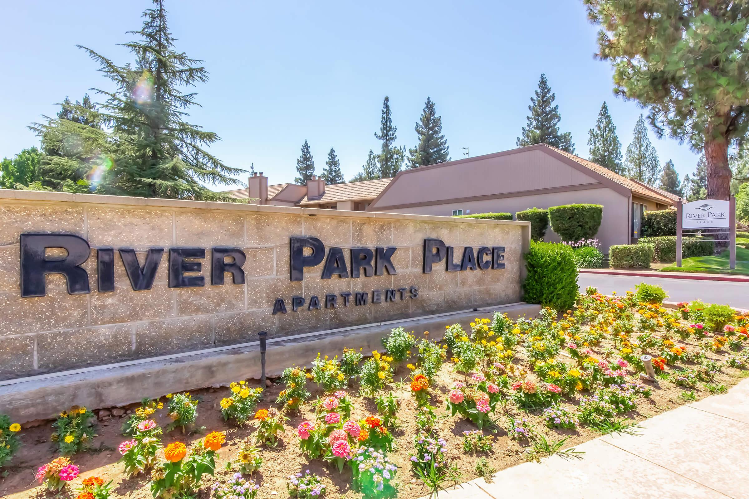 Sign for River Park Place Apartments with a landscaped flower bed in front, featuring colorful plants. In the background, a building and tall trees are visible against a clear blue sky.