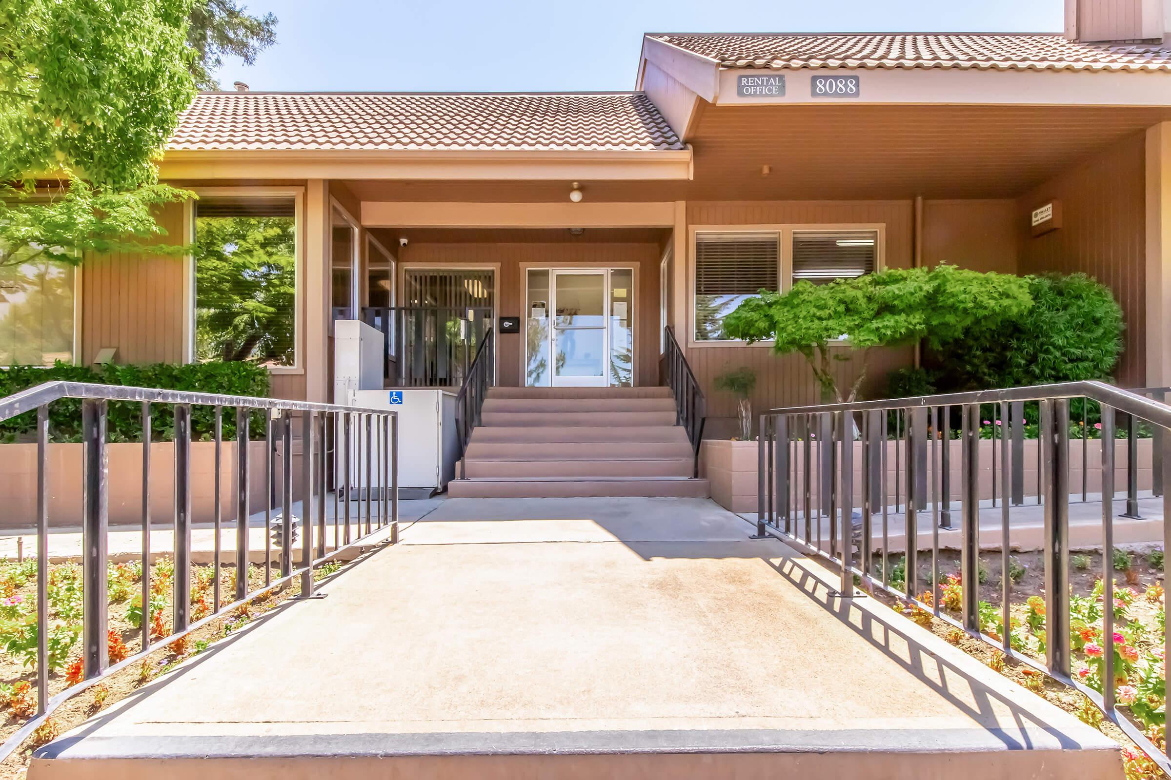 A modern rental office building with a welcoming entrance featuring a set of stairs, surrounded by landscaped greenery and colorful flowers. The exterior has a warm tone, and large windows provide a view into the building's interior. A sign indicates the building number (8088).