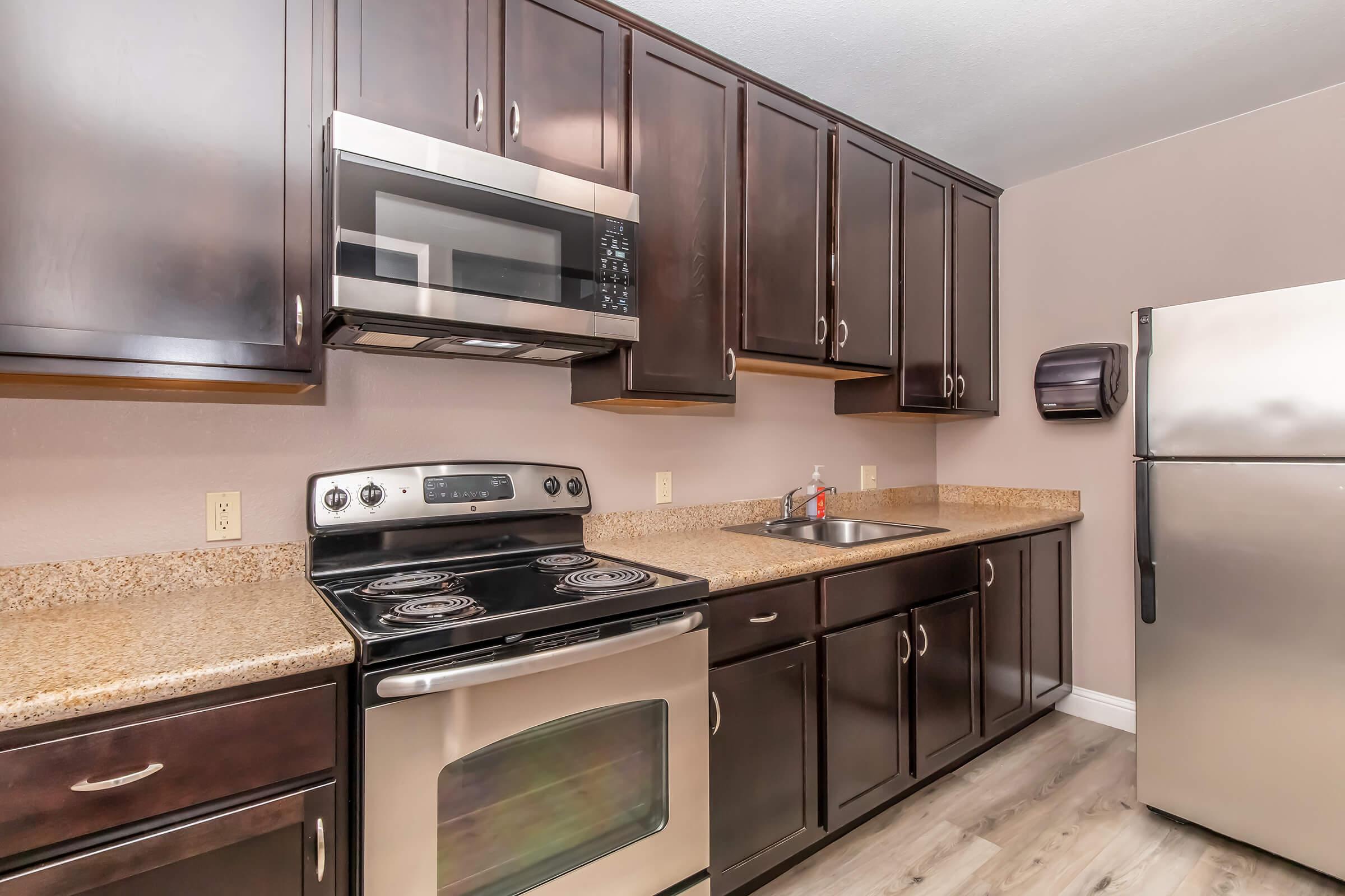 Modern kitchen featuring dark wood cabinets, stainless steel appliances including a microwave and refrigerator, a smooth-top stove, and a granite countertop. Natural light highlights the space, creating a warm and inviting atmosphere.