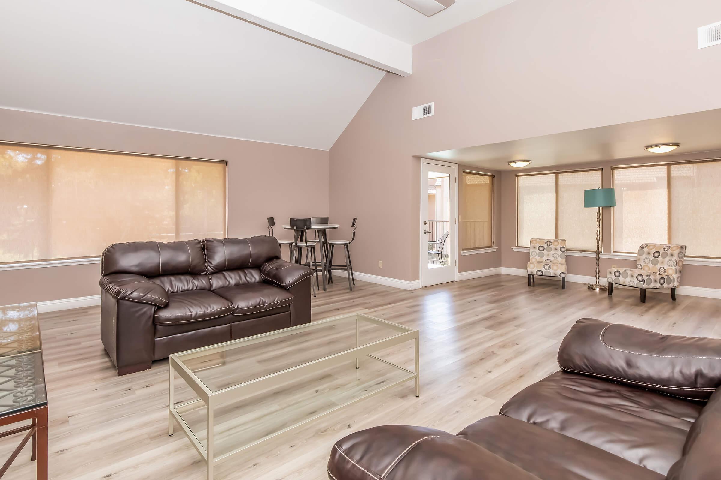 A spacious living room featuring two brown leather sofas, a glass coffee table, and large windows with shades. In the background, there are two stylish chairs and a bar-height table with stools. The room is well-lit with neutral-colored walls and a high ceiling, creating an inviting atmosphere.