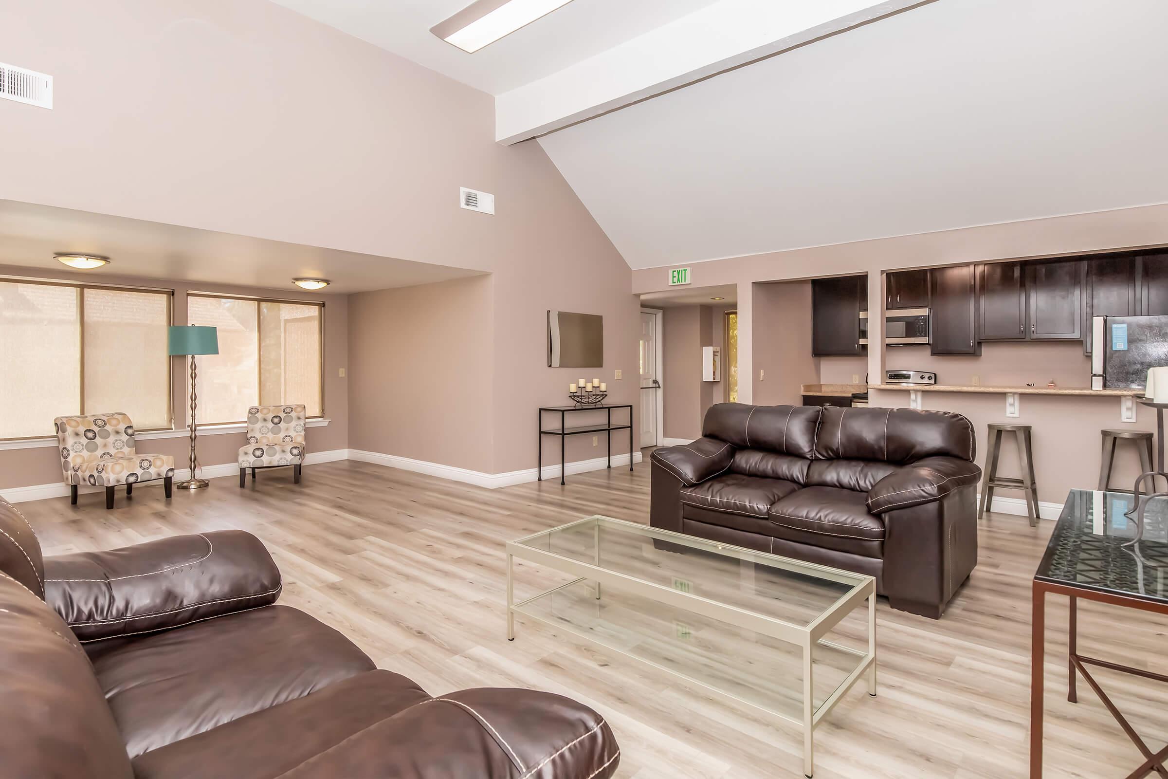 Spacious and modern living room with brown leather sofas, a glass coffee table, and two patterned chairs. The room features light-colored walls, large windows allowing natural light, and an open layout leading to a kitchen area with dark cabinetry. Brightly lit, the space feels inviting and contemporary.