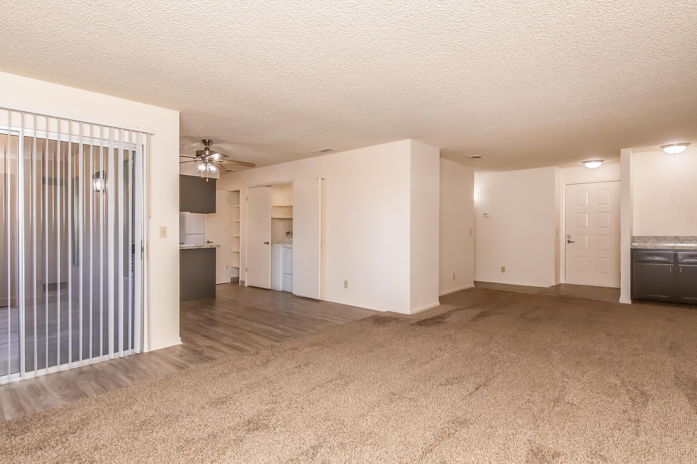 Spacious living area with carpeted flooring, featuring a ceiling fan, and an open layout leading to a kitchen area. Sliding glass doors provide access to an outdoor space. The kitchen shows visible cabinetry, and there's a light-colored wall with a door at the far end. Bright and airy atmosphere.
