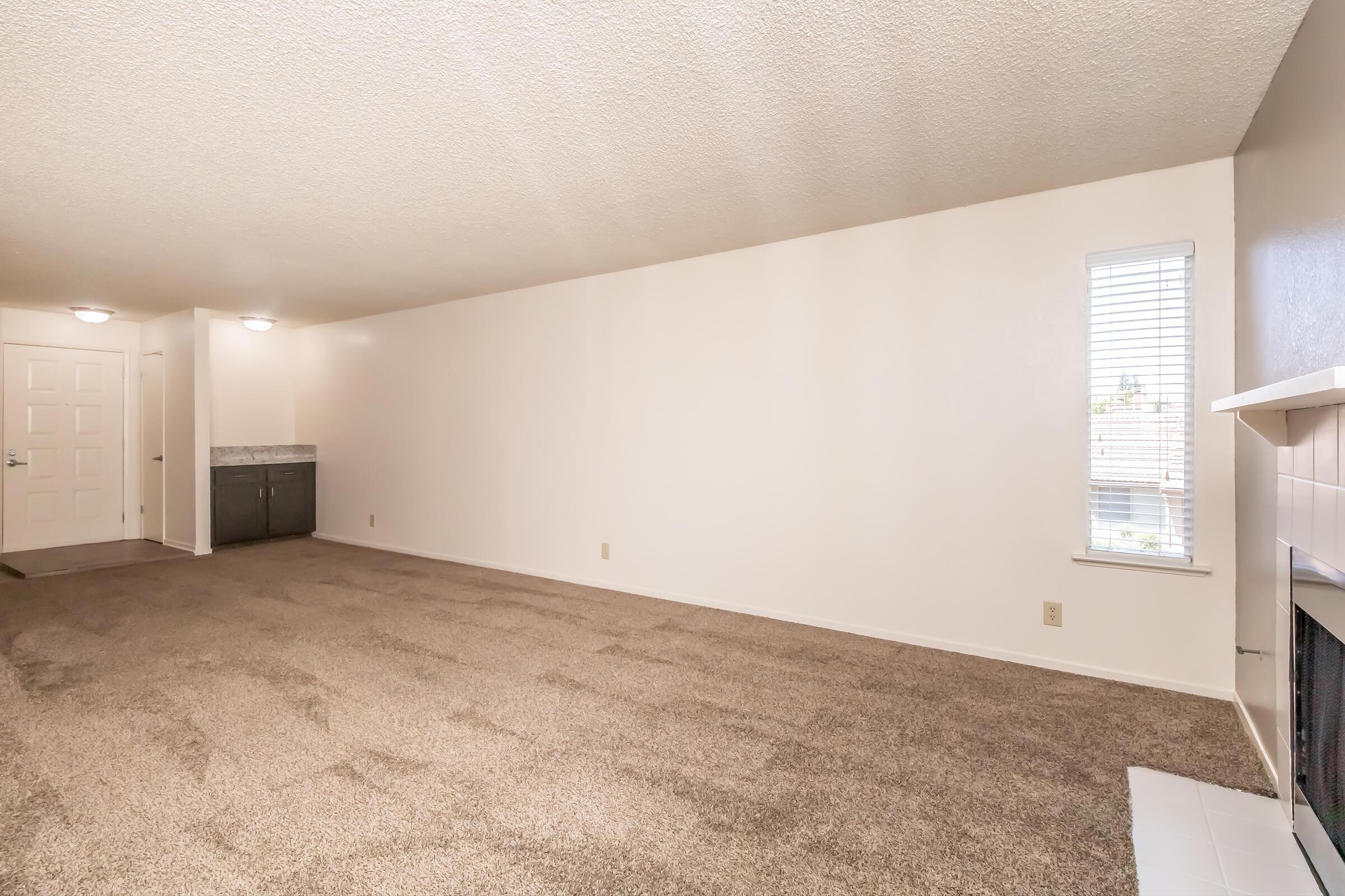 A spacious, empty room with beige carpet and light-colored walls. There is a window with blinds, allowing natural light to enter. On one side, a door leads to another area, while a small kitchenette is visible in the back. The room features a minimalist design, providing a blank canvas for furnishings.