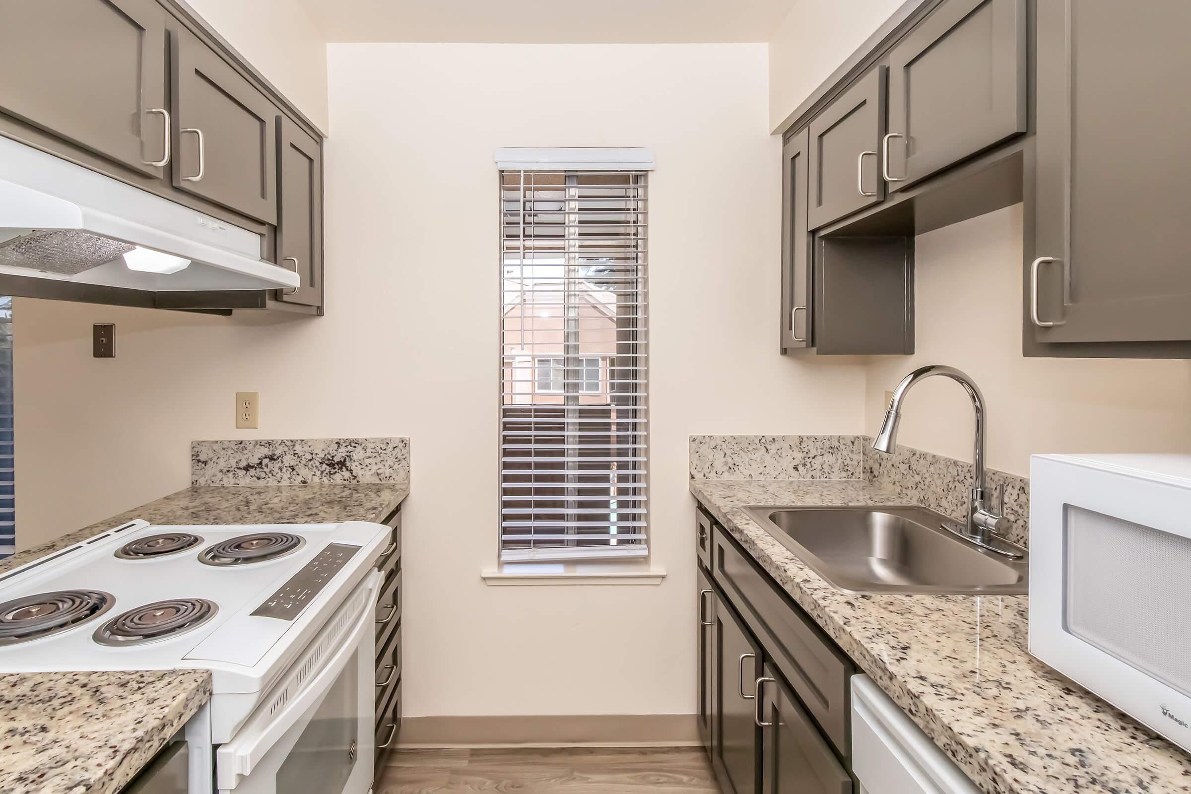 A modern kitchen featuring dark cabinets, a granite countertop, a stove with four burners, a sink, and a microwave. A window with blinds allows natural light in, and the flooring is a light wood finish. The compact layout includes essential kitchen appliances and storage.