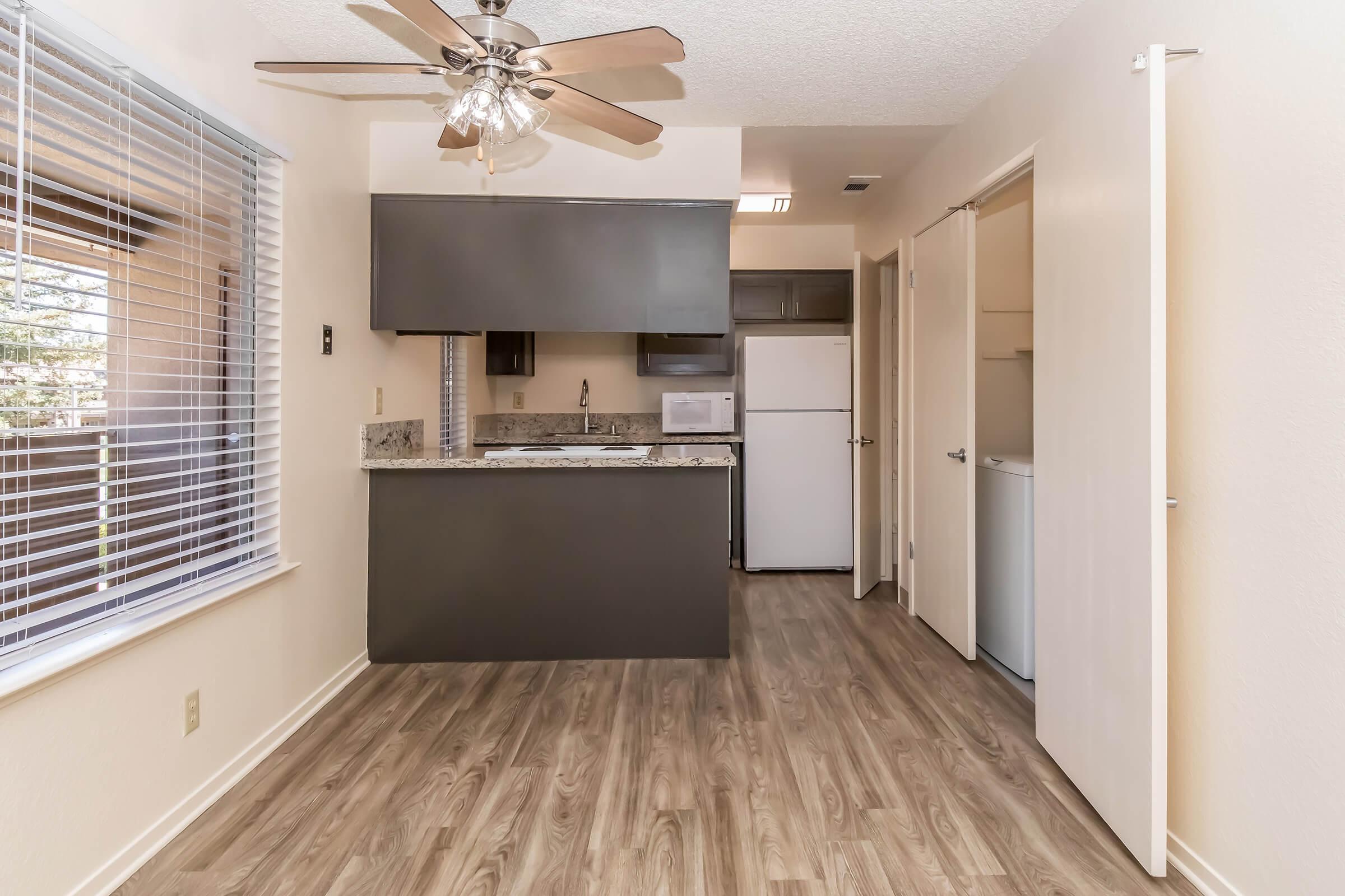 Interior view of a kitchen and dining area featuring laminate flooring, a ceiling fan, and an open design. The kitchen has dark cabinets, a granite countertop, and white appliances, including a refrigerator and microwave. Natural light enters through a window with blinds.