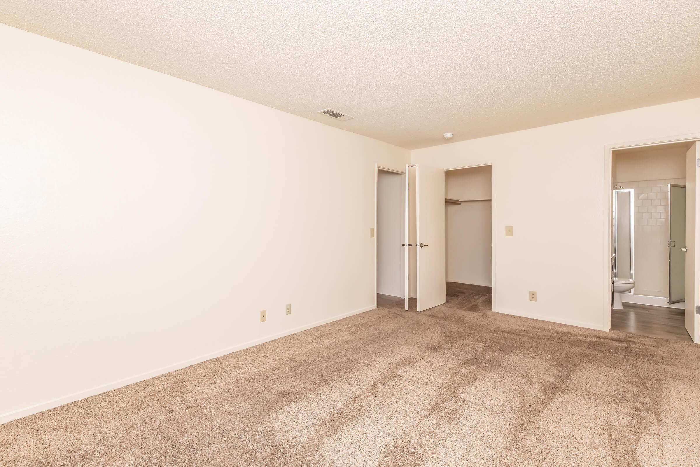 A spacious, empty room with light beige walls and carpet. There are two doorways leading to adjacent areas, one opening to a closet and the other to a bathroom. The room is well-lit with natural light and has a simple, clean look, making it ready for personalization.