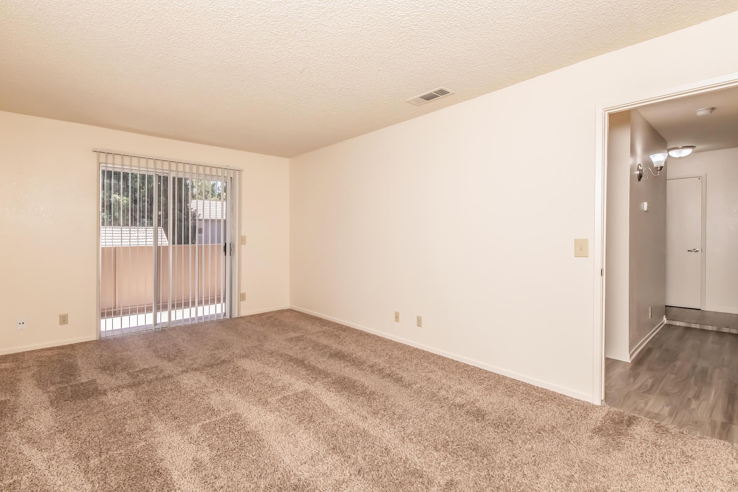 A spacious, empty living room featuring beige carpeting, light-colored walls, and a sliding glass door leading to an outdoor area. The room has no furniture and minimal decor, providing a blank canvas for personal styling. A doorway on the right leads to another part of the space.