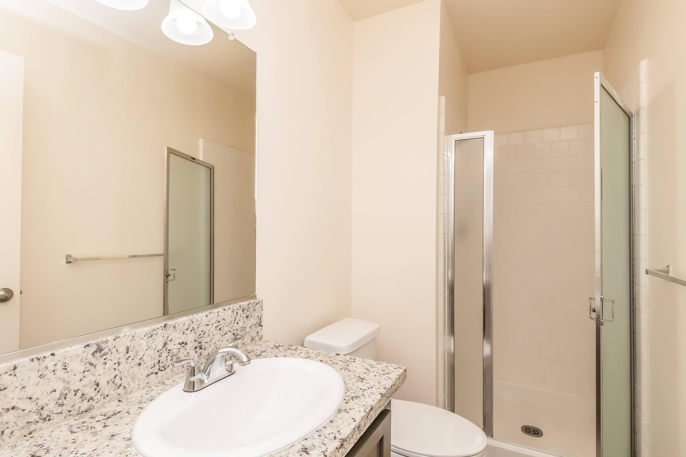 A clean bathroom featuring a white sink with a granite countertop, a mirror, a toilet, and a glass-enclosed shower. The walls are light-colored, and there is a towel bar on one side. Overall, the space appears modern and well-maintained.