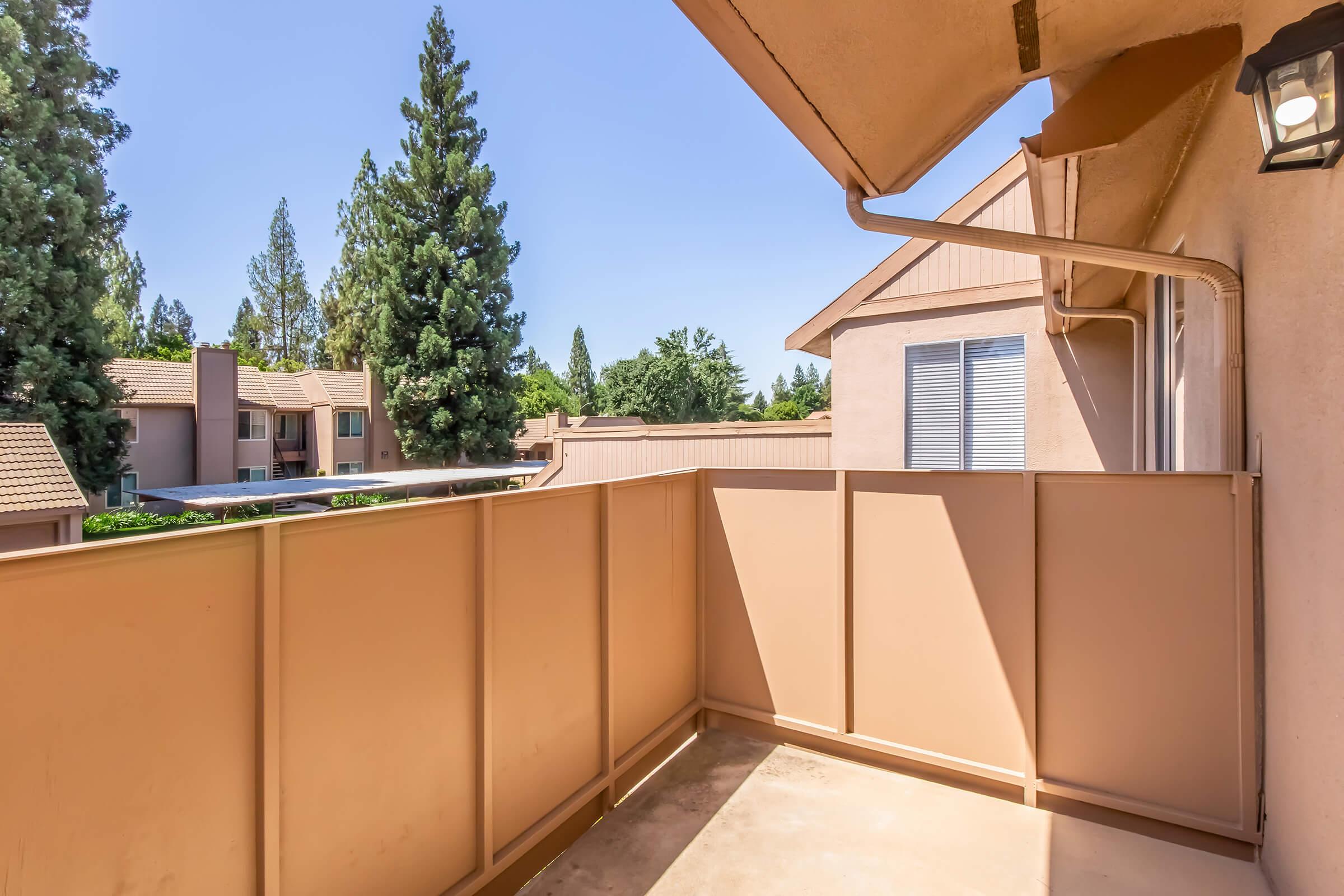 A clear blue sky above a small balcony with a tan-colored railing. In the background, tall green trees and a few residential buildings are visible, creating a serene outdoor setting. The balcony floor is empty, and sunlight brightens the area.