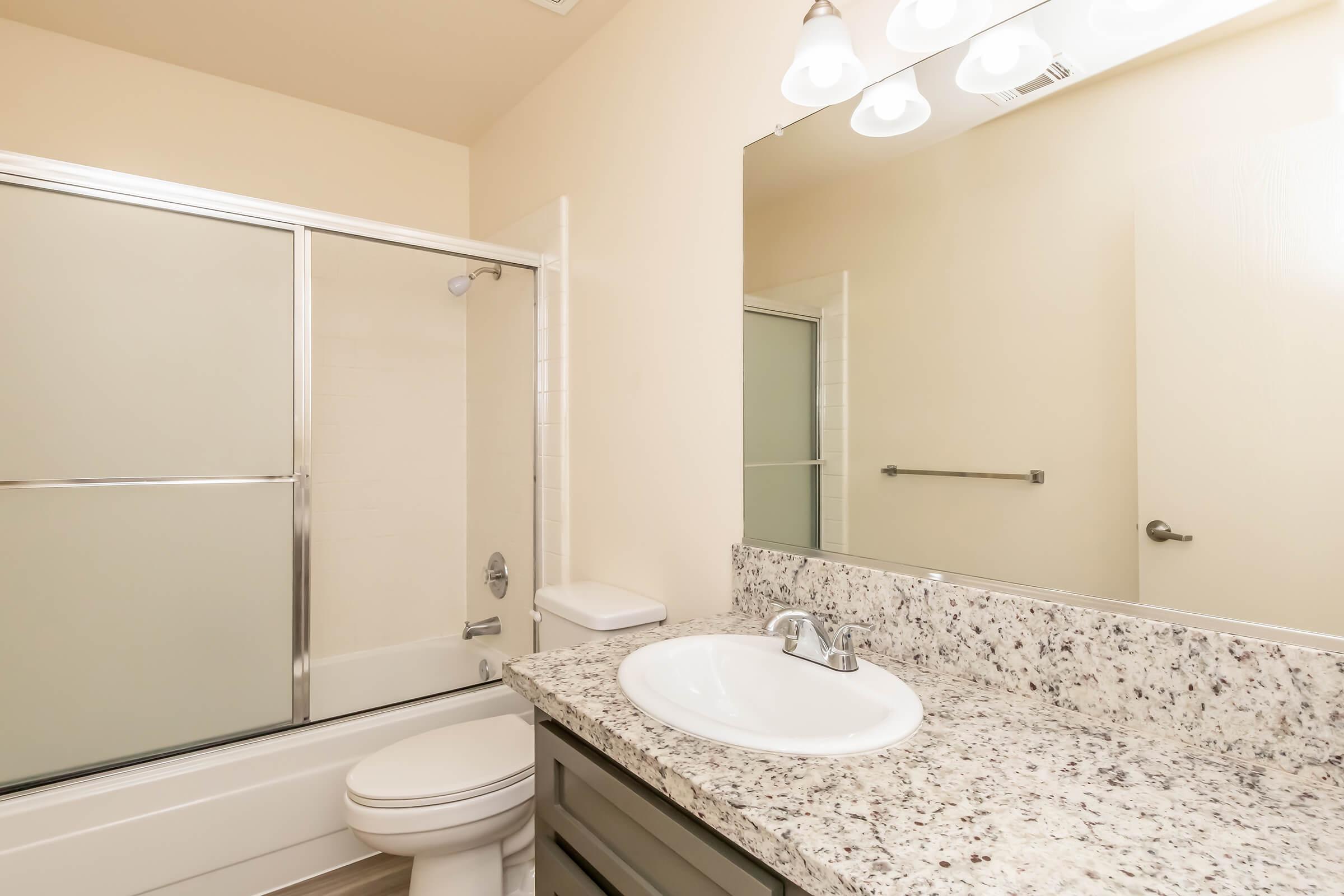 A clean and modern bathroom featuring a glass shower enclosure, a white bathtub, a granite countertop with a sink, and a large mirror. The walls are painted in a soft beige color, and the lighting is bright from three overhead light fixtures.