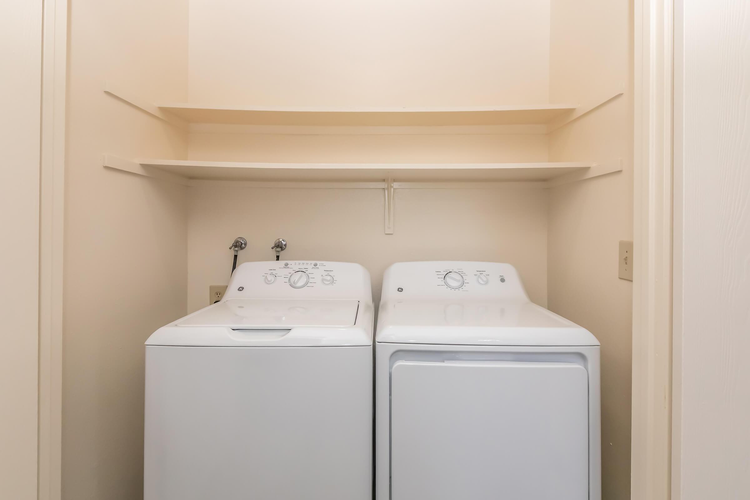 A clean, organized laundry room featuring a white washing machine and a white dryer side by side, placed against a light-colored wall. Above them, there are empty shelves for storage. The space is well-lit and minimalistic, emphasizing functionality.