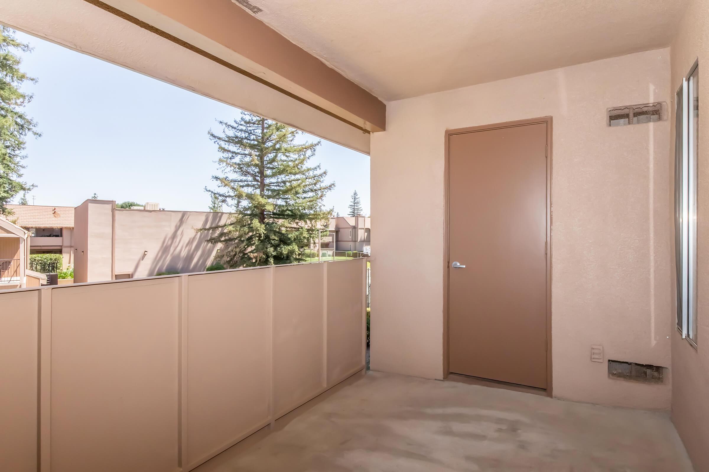 A simple, empty balcony with a tan railing and a closed brown door. The view includes trees and nearby buildings under a clear blue sky. The floor is bare and the walls are light-colored.
