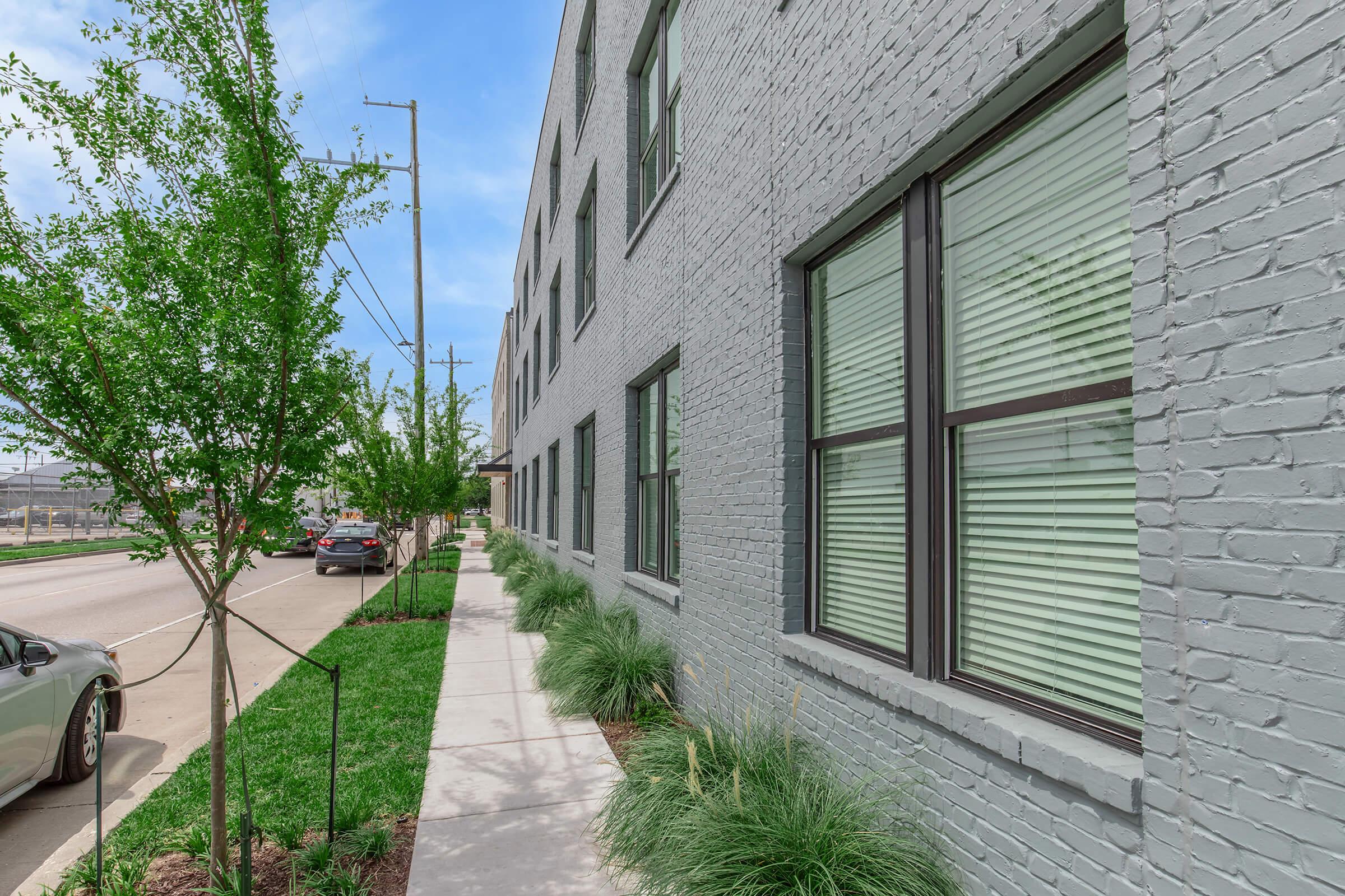 A sidewalk lined with green plants leads to a gray brick building with large windows featuring blinds. The street is visible on the left, with parked cars and electrical poles nearby, under a clear blue sky.