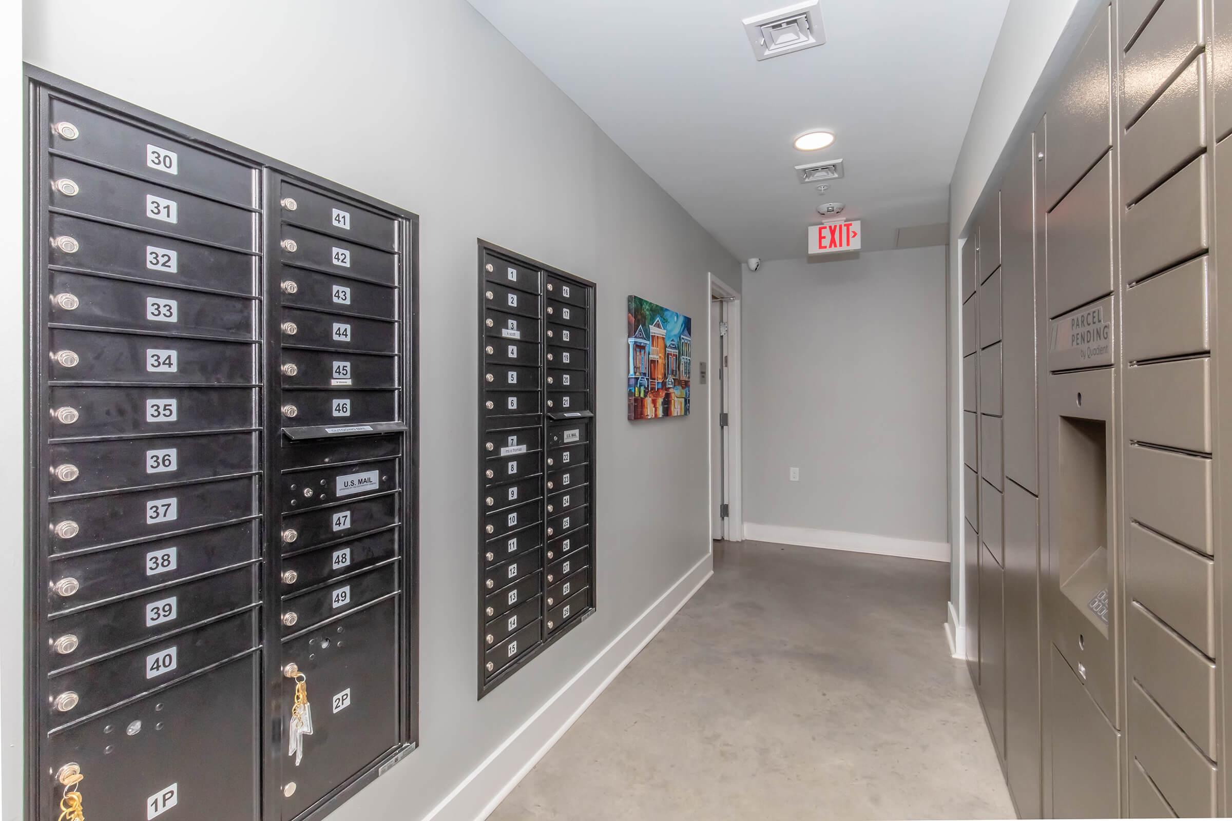 A narrow hallway featuring several black mailboxes on the left side, a single black mailbox unit on the right, and a colorful framed painting on the wall. The floor is concrete, and there's an exit sign visible at the end of the corridor.