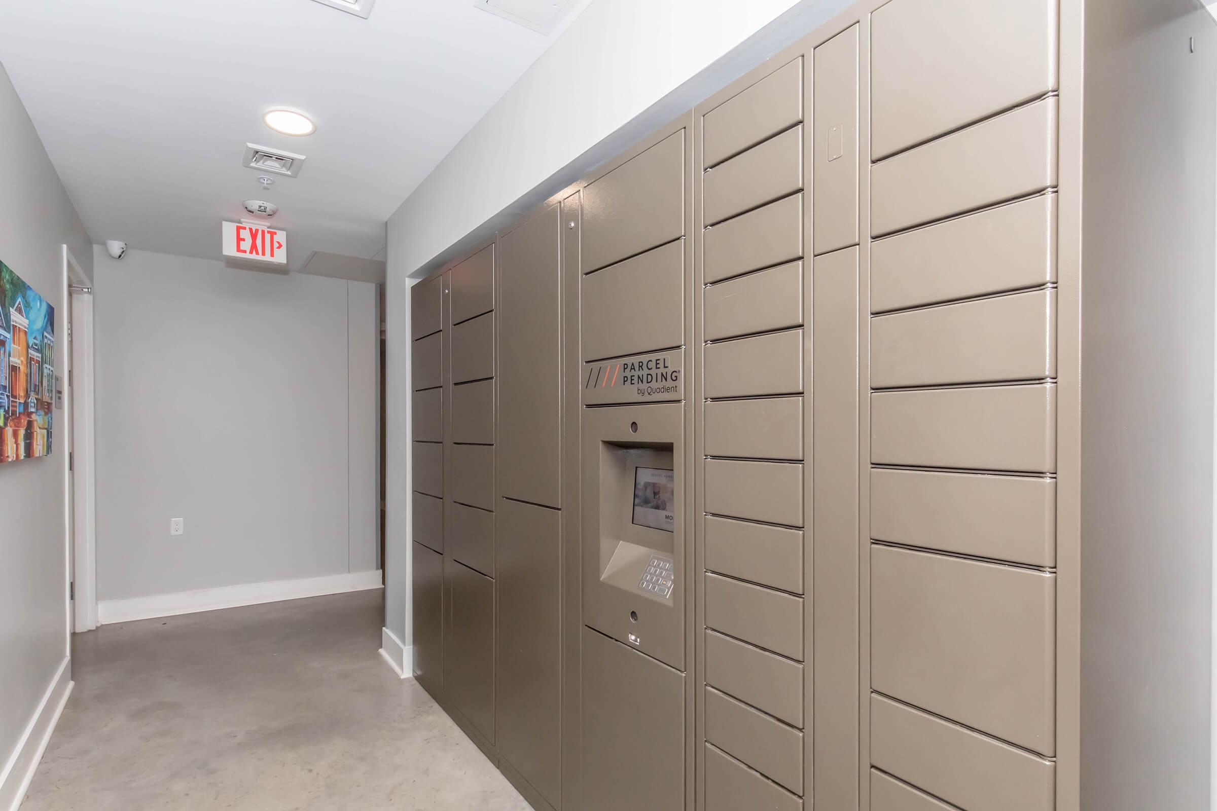 Interior hallway featuring a series of beige parcel lockers along one wall. A small electronic kiosk is positioned next to the lockers, which is used for package retrieval. The floor is concrete, and exit signage is visible at the end of the hallway.