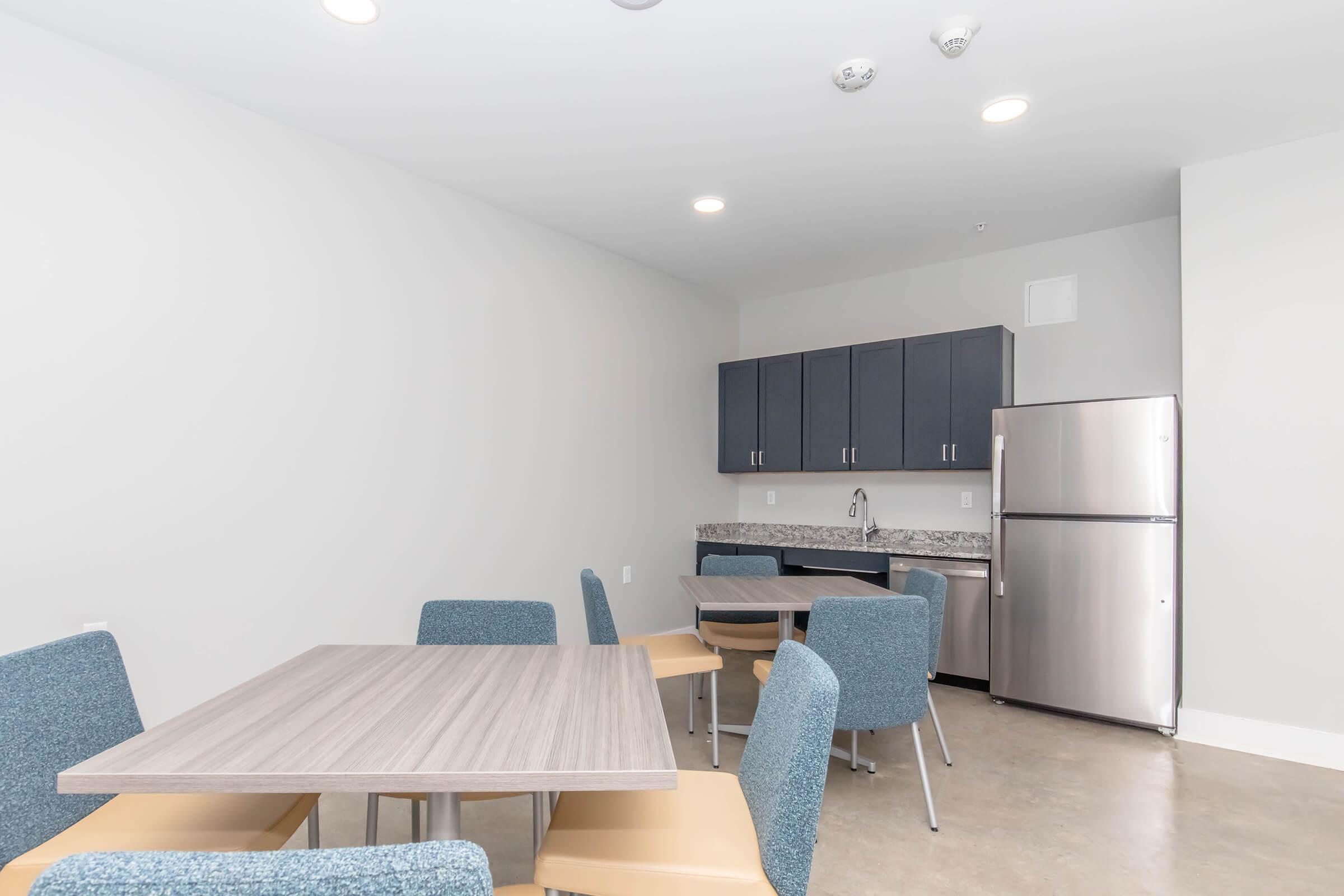 A modern kitchen area featuring a gray tabletop with beige and blue chairs. The space includes dark blue cabinets, a stainless steel refrigerator, and a countertop. Bright lighting enhances the open, minimalist design with light-colored walls and a polished concrete floor.