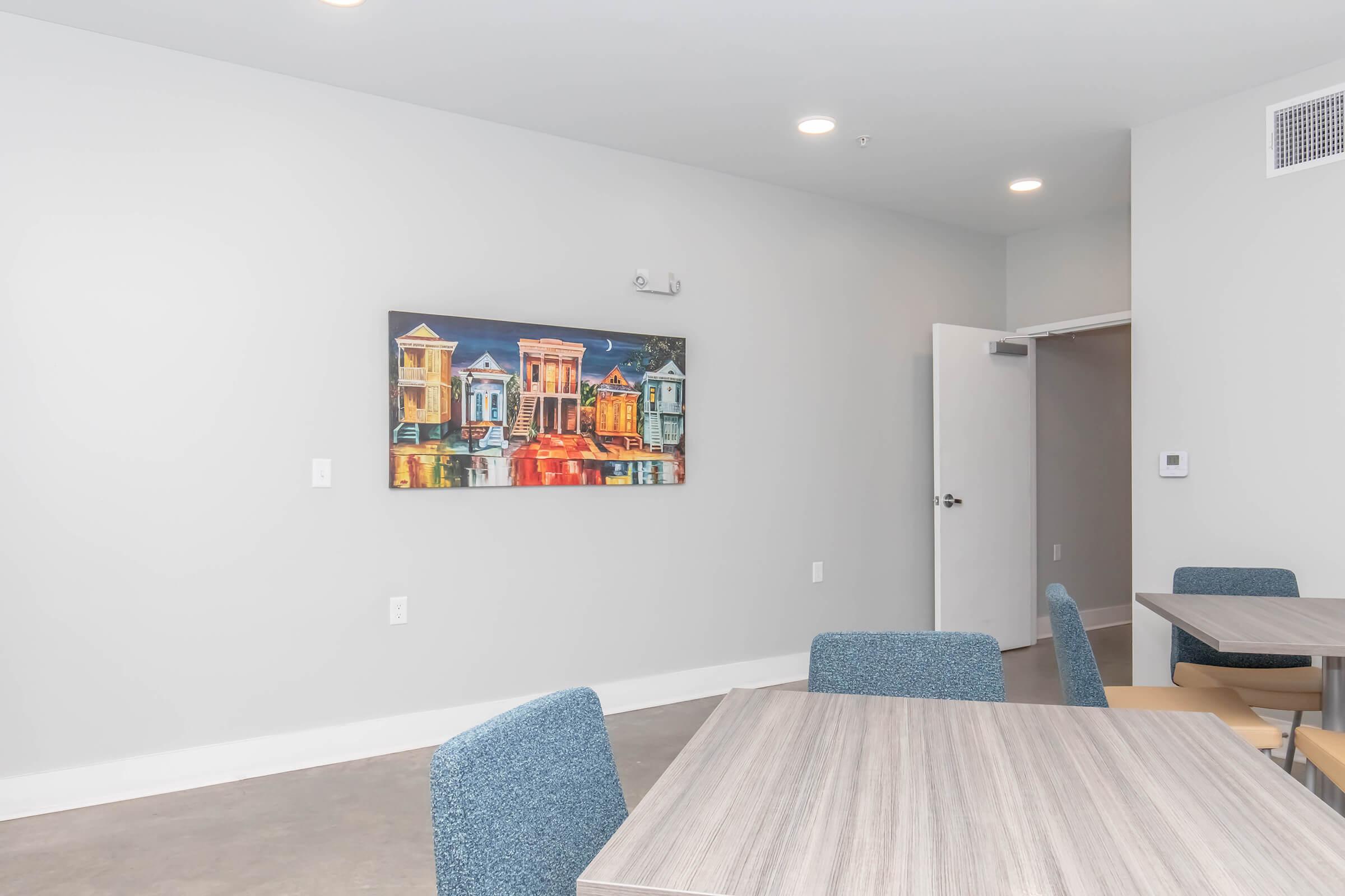 A modern interior space featuring a light gray wall with a colorful painting of houses hanging on it. In the foreground, there are wooden tables and blue upholstered chairs. A door is partially open, leading to another room. The overall atmosphere is bright and contemporary.