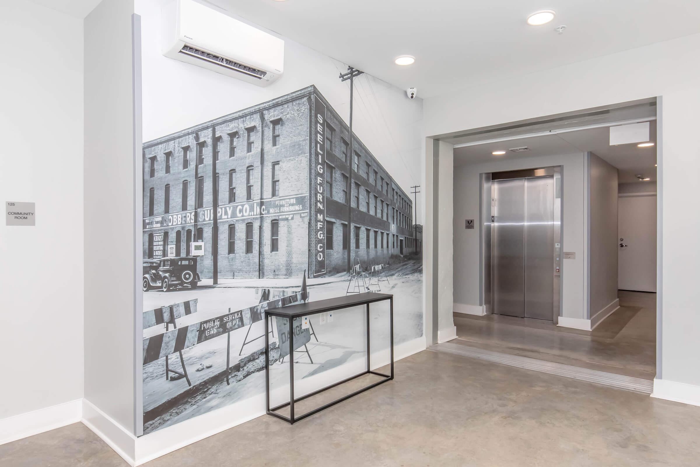 A monochrome mural of a historical building on a wall, featuring a vintage truck outside. In the foreground, there's a modern black table and an elevator entrance. The interior has a contemporary design with concrete flooring and neutral-colored walls.