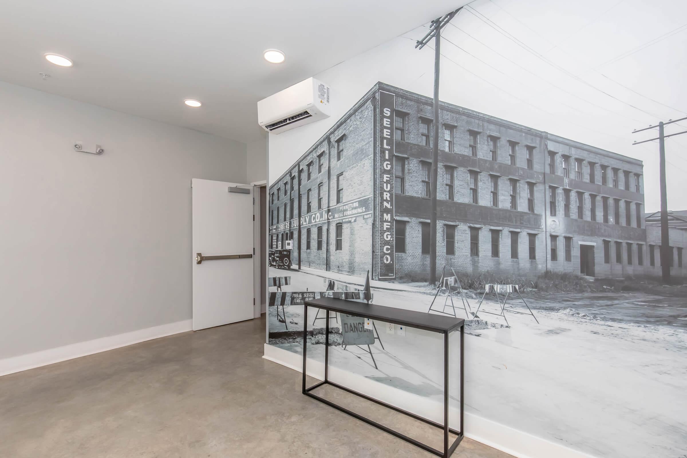 An interior space with a concrete floor and a modern metal table against a black-and-white mural of a historic brick building. The wall feature includes light fixtures on the ceiling and a door leading to another room. The mural adds an architectural element to the contemporary design.