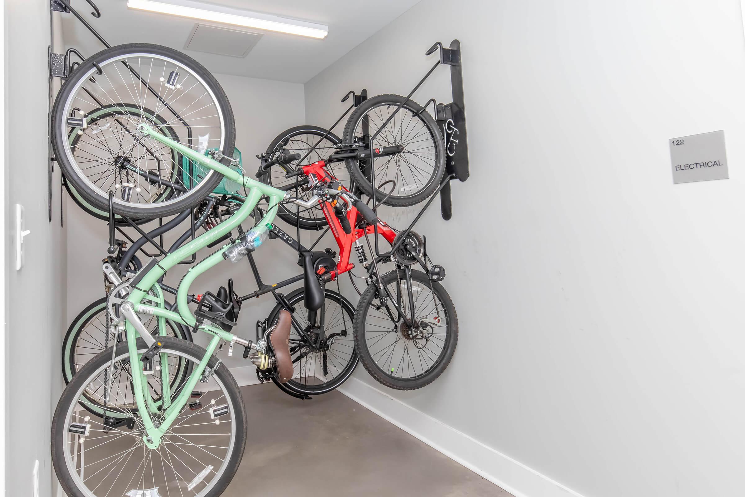 A narrow room displaying several bicycles neatly hung on wall-mounted racks. The bikes include a mint green bicycle, a red mountain bike, and others in various colors, all arranged efficiently to save space. A sign on the wall indicates the room number and designates it as "Electrical."