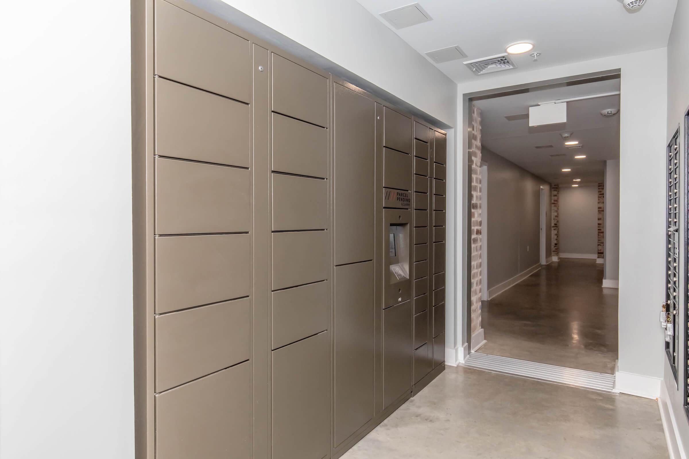 A modern hallway featuring beige storage lockers along one wall. The lockers are designed for package delivery and retrieval. The space has a polished concrete floor and white walls, and a narrow corridor leads further into the background, with dim lighting and a minimalist aesthetic.
