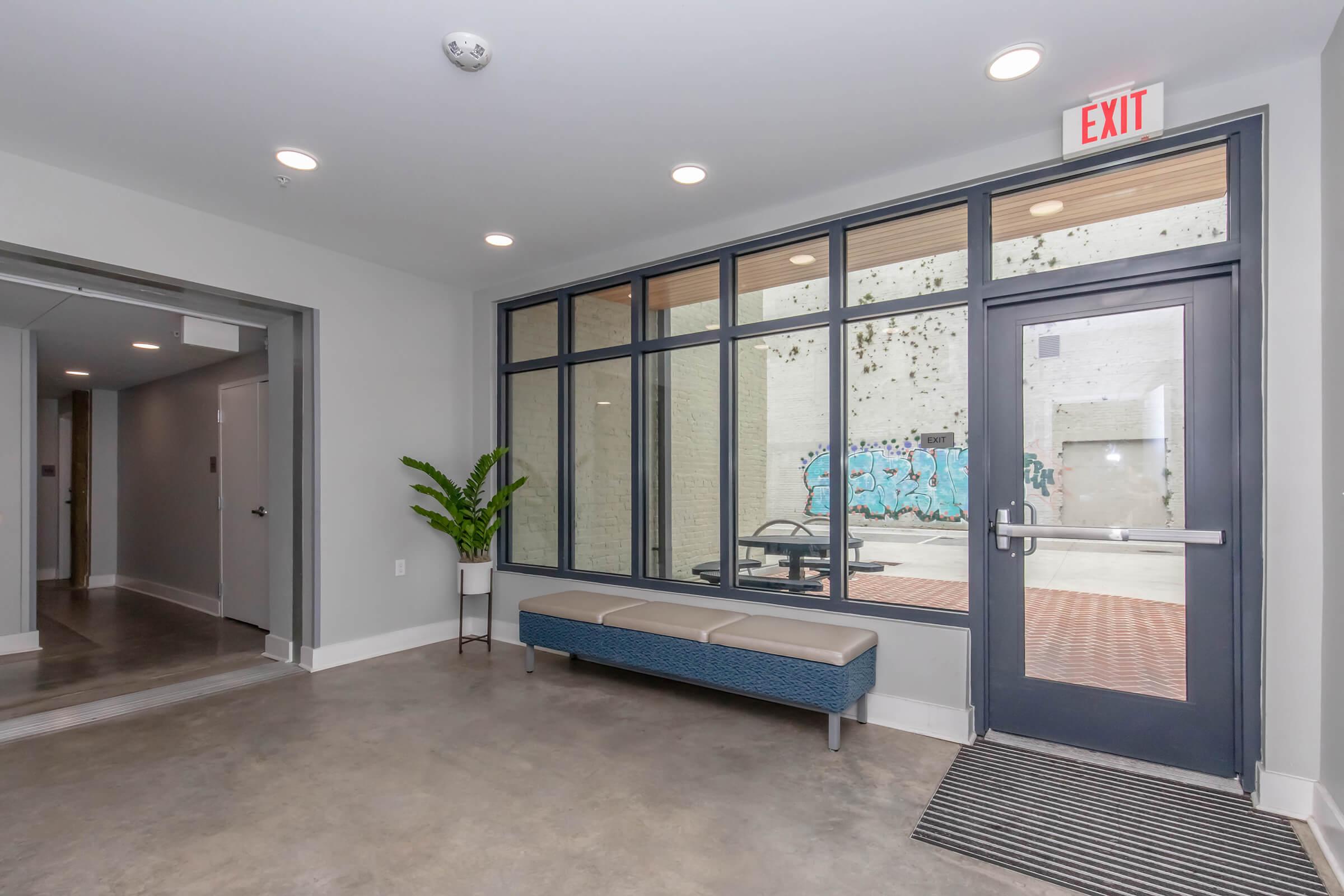 A modern lobby area with a large window showcasing an outdoor space featuring graffiti. There is a bench against the wall and a potted plant nearby. An elevator door is visible to the left, and an exit sign indicates the way out. The flooring is polished concrete, creating a sleek, contemporary look.