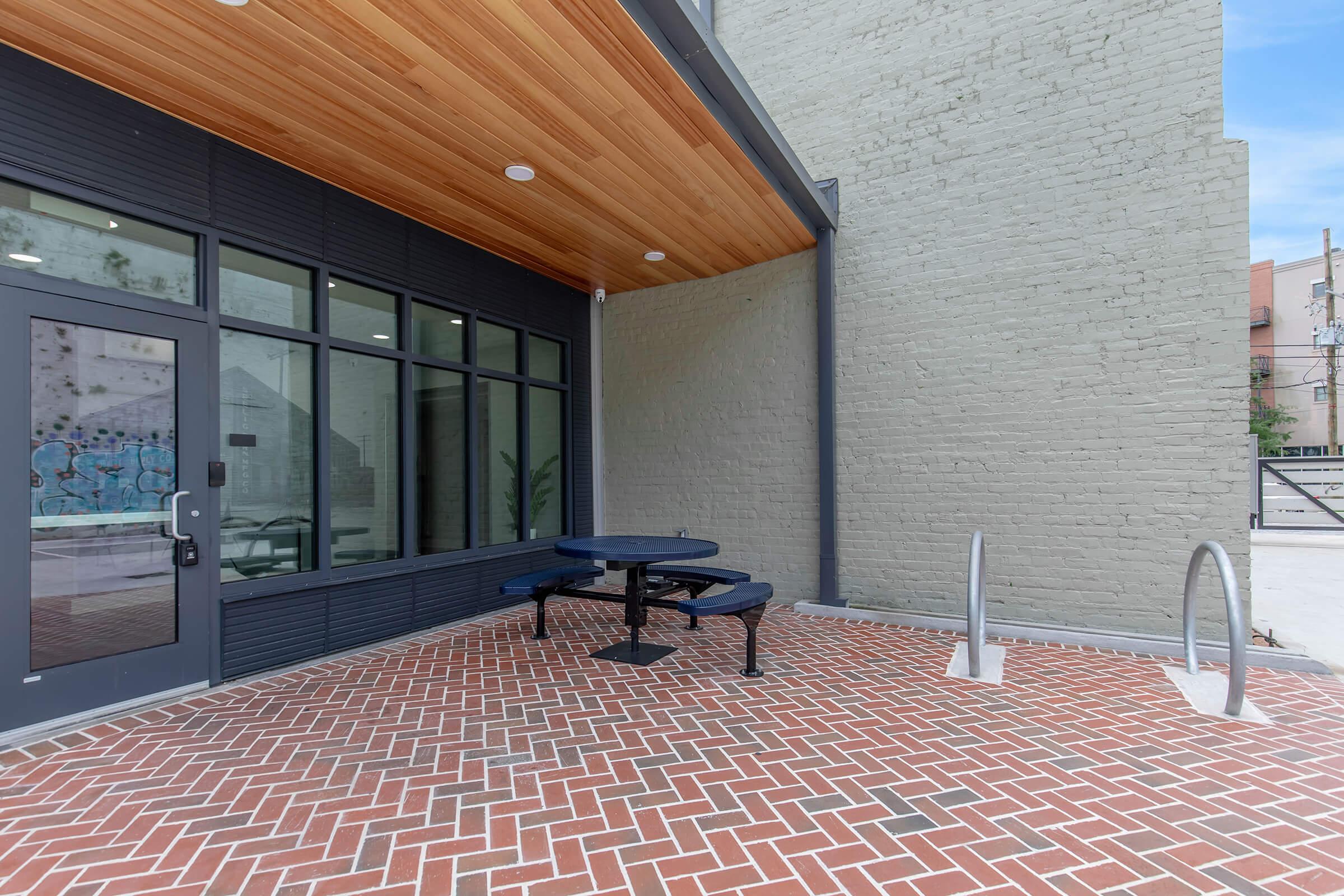A modern outdoor space featuring a brick patio with a black picnic table, surrounded by large windows of a building. The area is under a wooden overhang, with a bicycle rack visible nearby, and graffiti art faintly visible on the adjacent wall. Bright blue sky in the background.