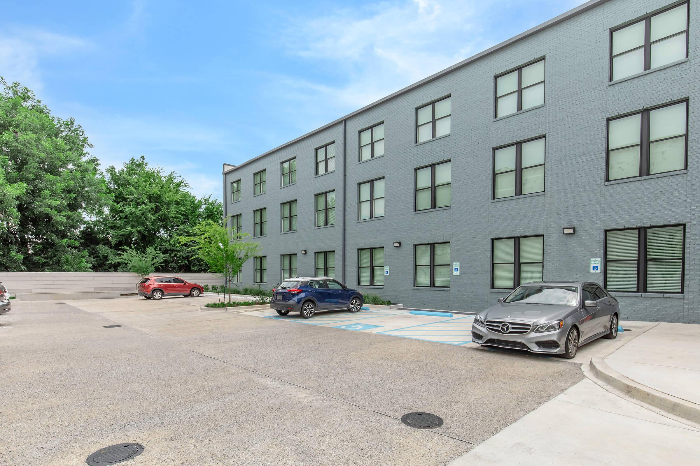A modern gray building with multiple windows on one side, accompanied by a parking lot containing a few parked cars. Lush green trees are visible in the background, adding to the overall serene atmosphere of the area. The scene is bright and well-lit, suggesting a pleasant day.