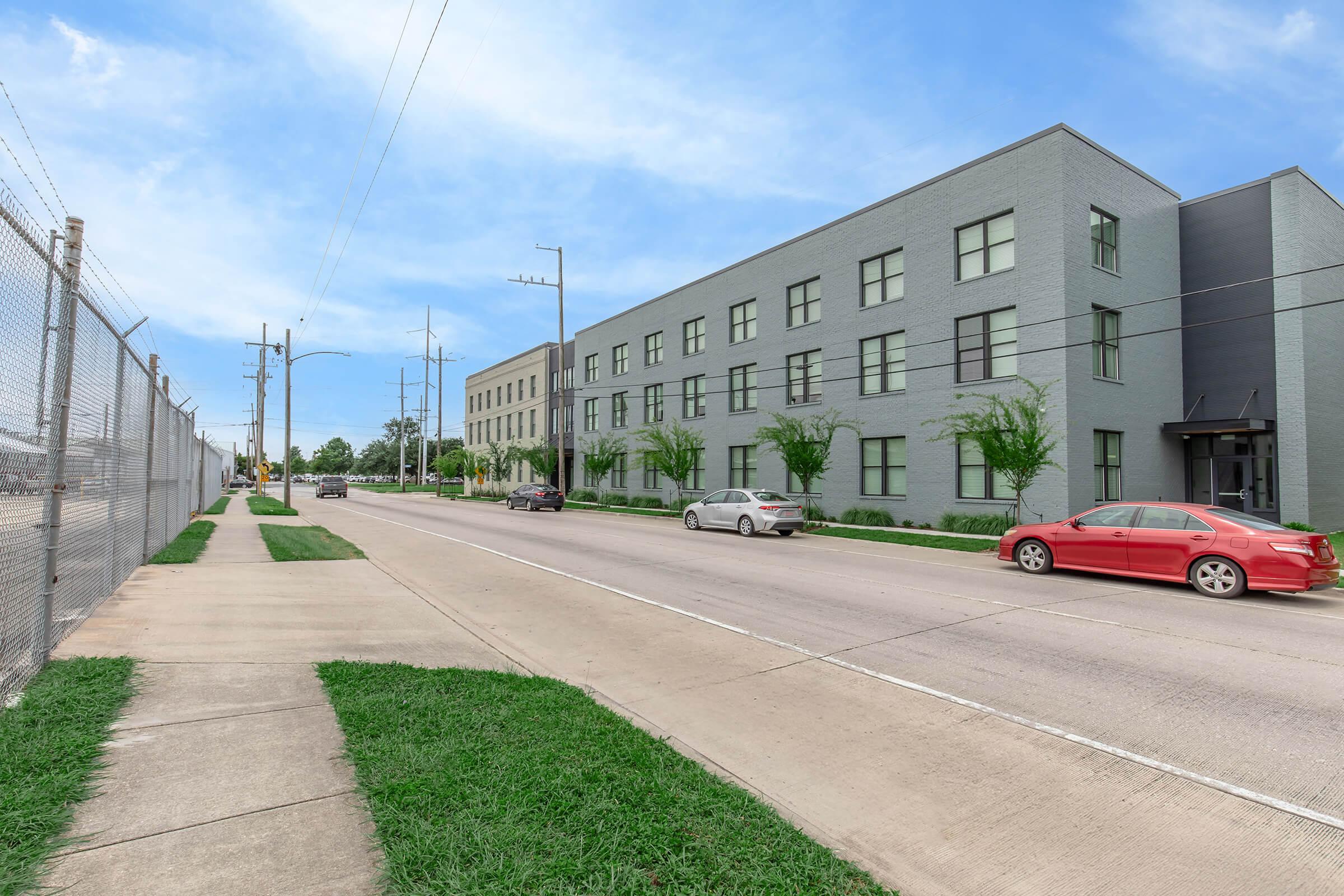 A street view of a modern, gray building with large windows, situated alongside a sidewalk. The scene features a well-maintained grassy area and trees, with light traffic of parked cars and a vehicle driving along the road. Power lines are visible in the background under a blue sky with scattered clouds.