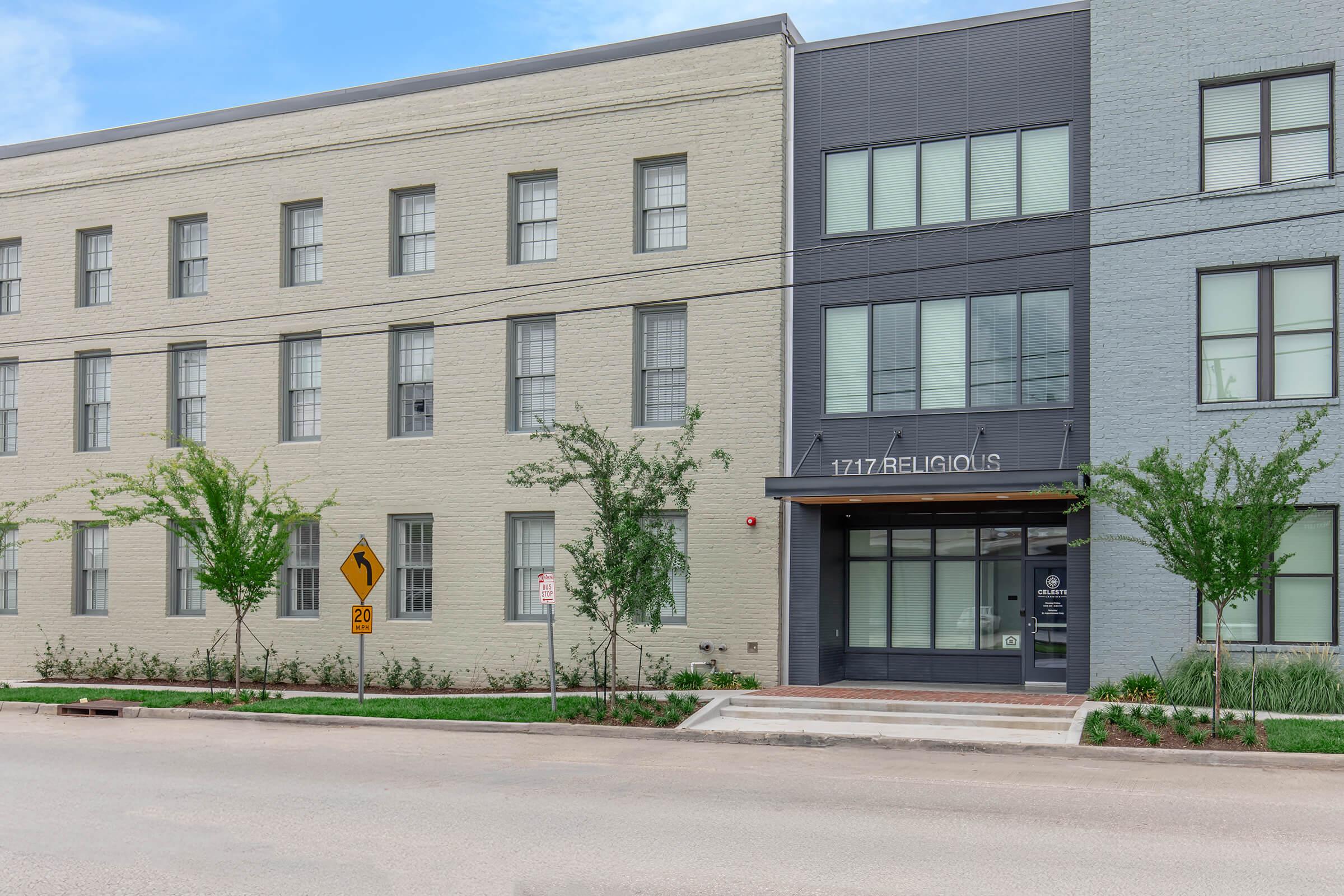 A modern building facade featuring two-tone brick exteriors with a gray and beige color scheme. The entrance is marked with the address "1717 Religious." Surrounding the building are small trees and planted areas, while a street sign indicates a curve ahead. The atmosphere is calm and urban.