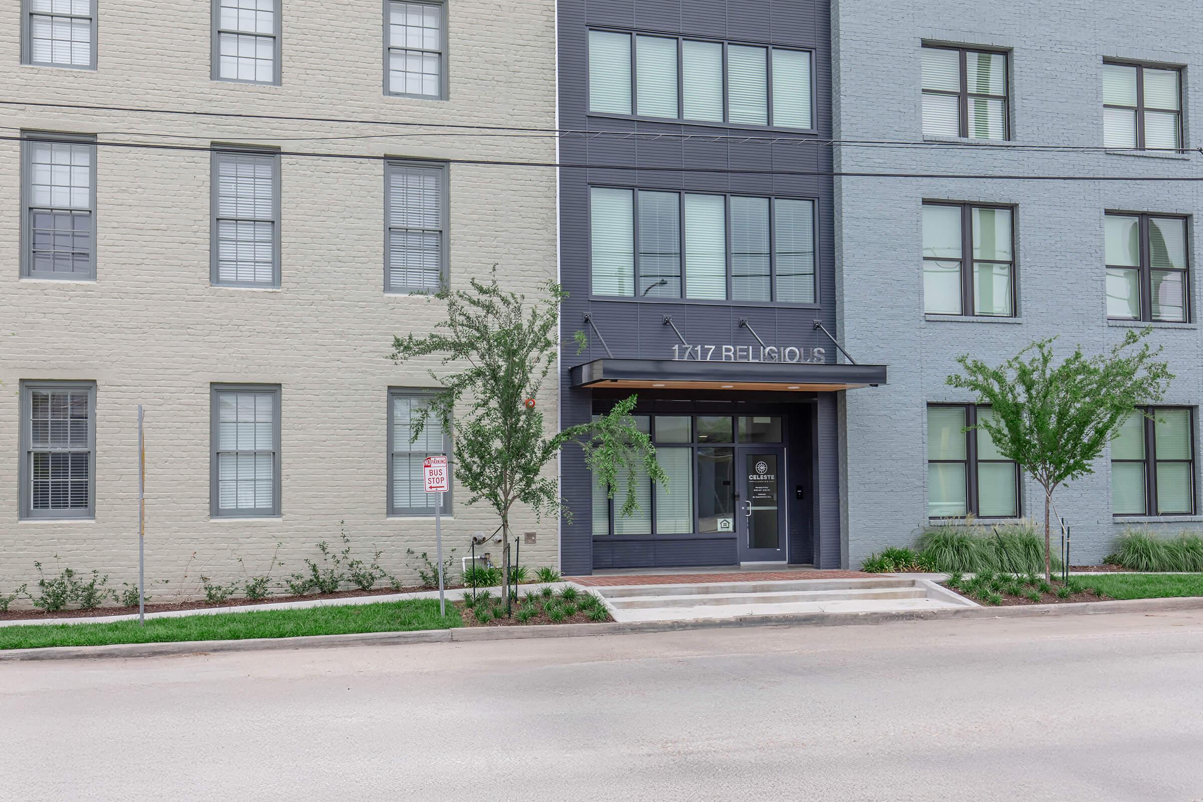 A modern two-story building with light-colored brick on the left and a darker facade on the right. The entrance features a canopy and large glass doors, flanked by small trees and landscaped greenery. The address, "1717 Religious St," is prominently displayed above the entrance.