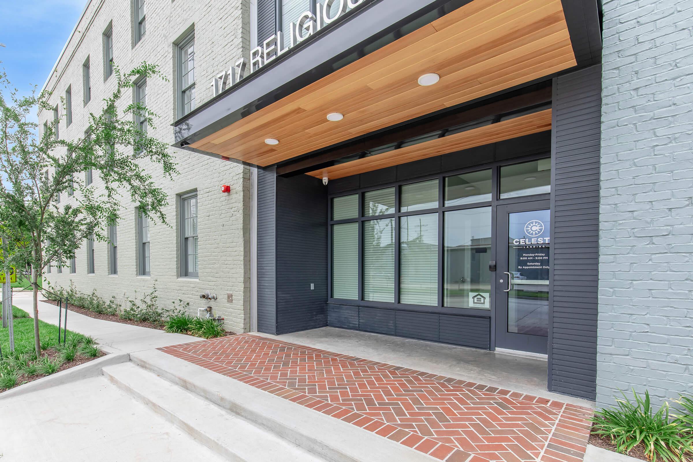 Exterior view of a modern building entrance, featuring a wood-paneled overhang, large glass windows, and a brick walkway. The entrance includes a glass door with a logo. Surrounding the entrance are manicured lawns and small trees, creating a welcoming atmosphere.