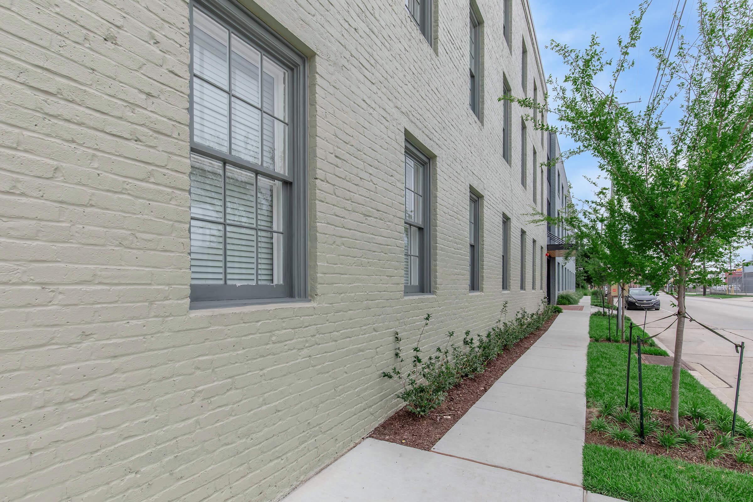 A street view of a modern building with a light green brick exterior, featuring large windows with white shutters. A clean sidewalk runs alongside, bordered by small shrubs and grass. Tree branches can be seen in the foreground, and a street is visible in the background under a clear blue sky.