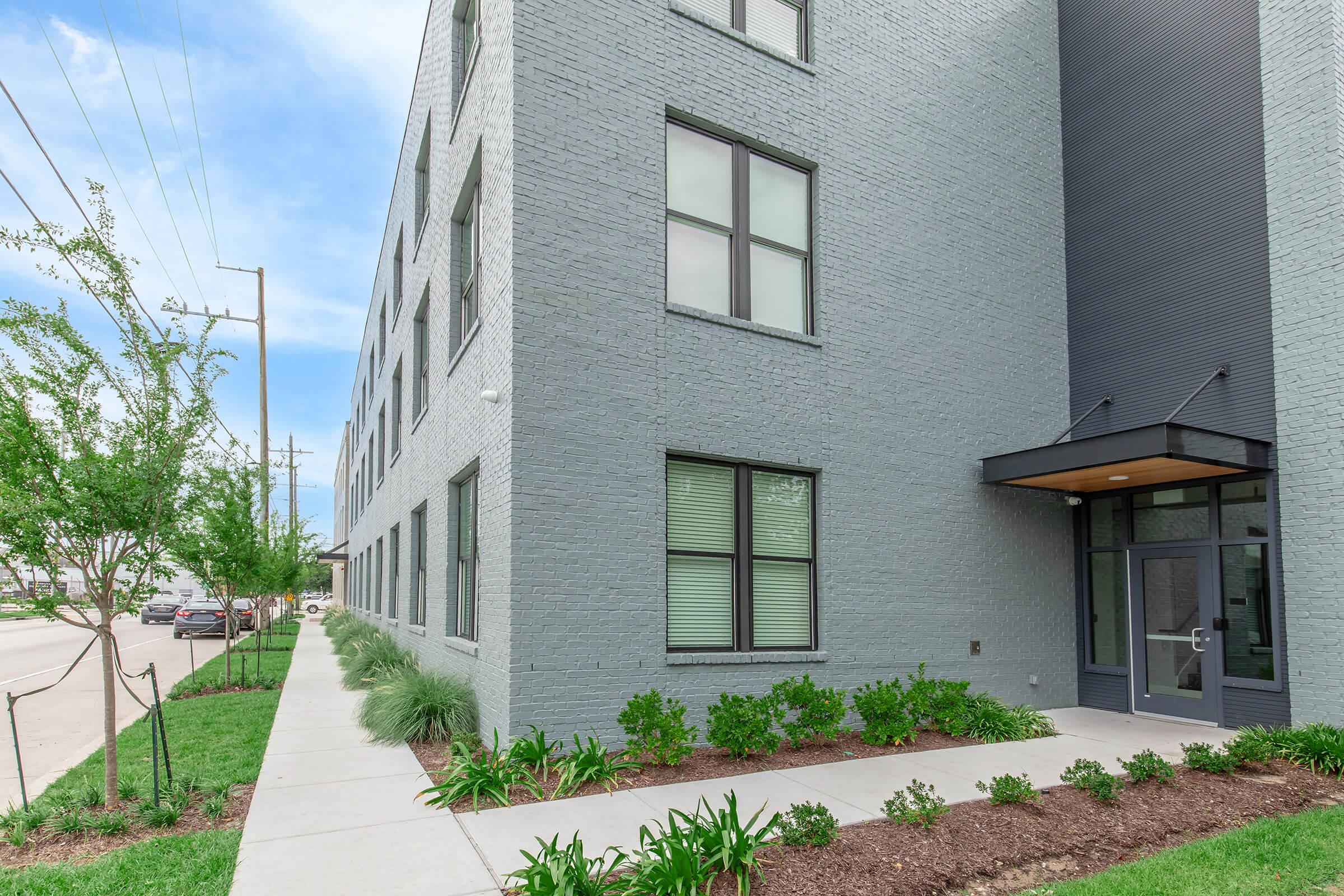 A modern gray building with large windows, featuring a flat-roof entrance and a landscaped area with green plants and freshly trimmed grass. Sidewalks run along the building, and a street with parked cars is visible in the background under a partly cloudy sky.