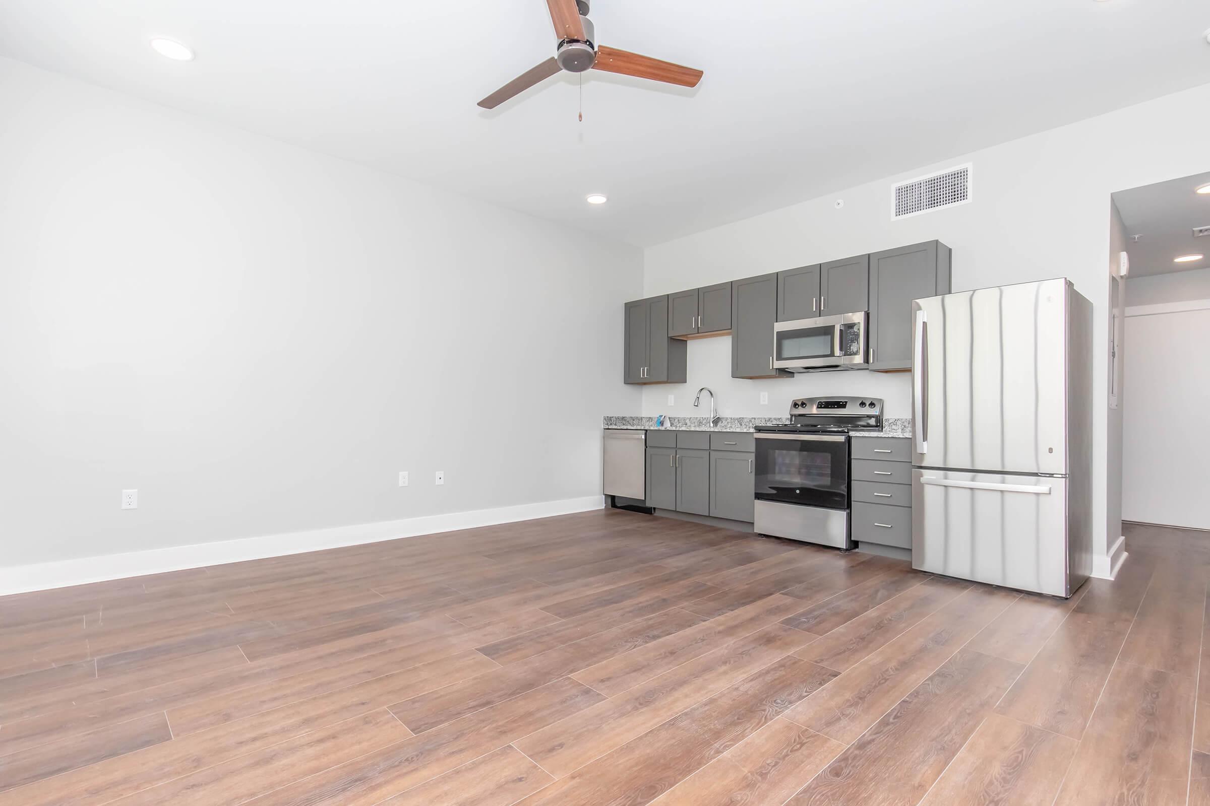 Modern kitchen with gray cabinets, stainless steel appliances, and a granite countertop. The open layout features wooden flooring and a ceiling fan. Natural light fills the space from windows, creating a bright and inviting atmosphere.