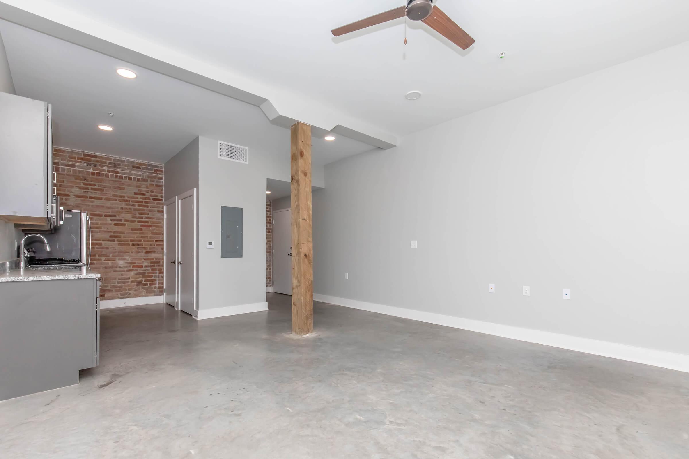 A spacious, modern interior of an apartment featuring a concrete floor, exposed brick wall, and a wooden support column. The kitchen area with stainless steel appliances is partially visible on the left. The walls are painted in a light gray color, and there is a ceiling fan above.