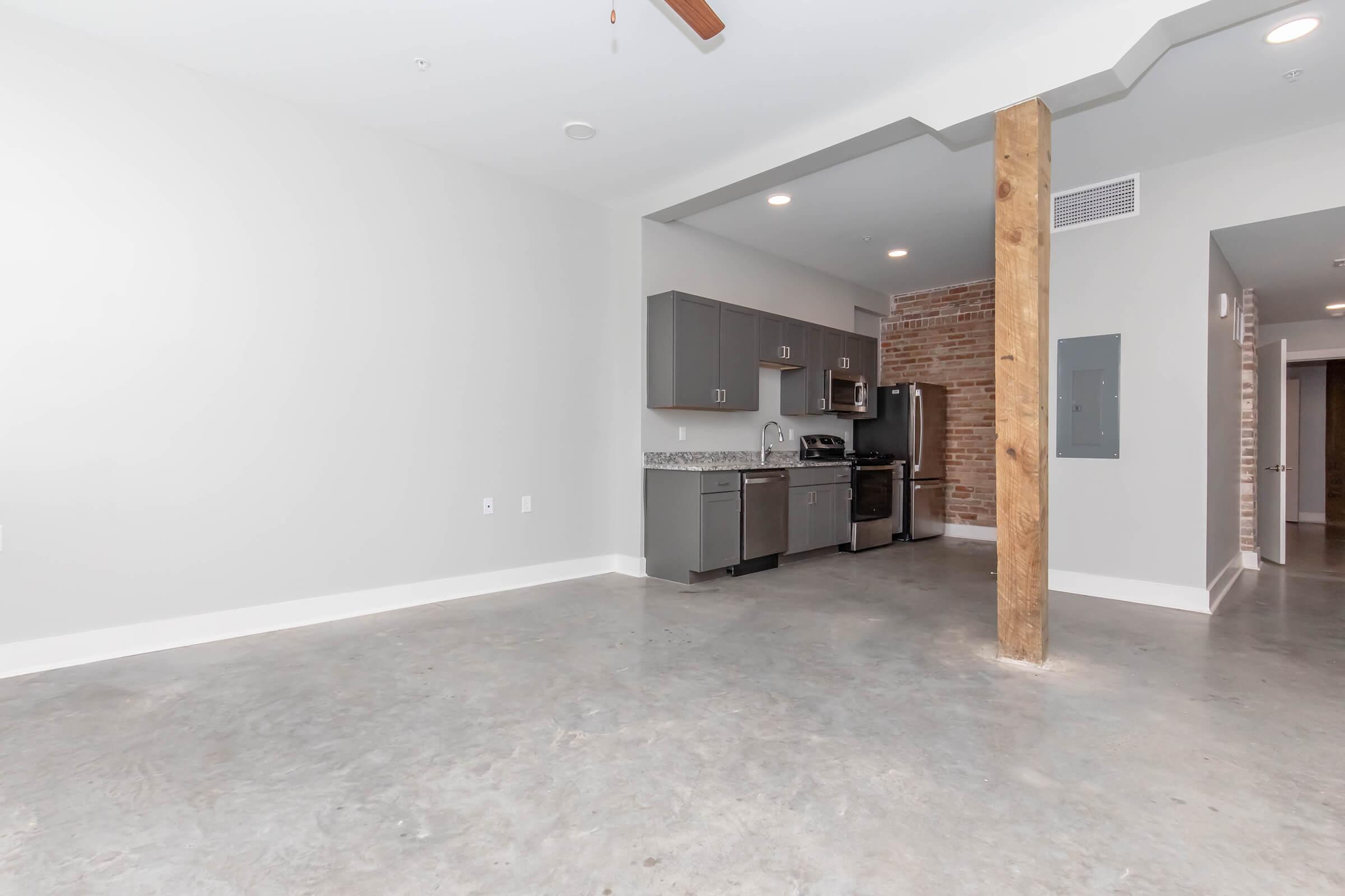 Spacious modern kitchen in a minimalist apartment featuring gray cabinetry, stainless steel appliances, and exposed brick accents. The open floor plan includes concrete flooring, a ceiling fan, and a wooden support beam. Natural light fills the room from a nearby window.