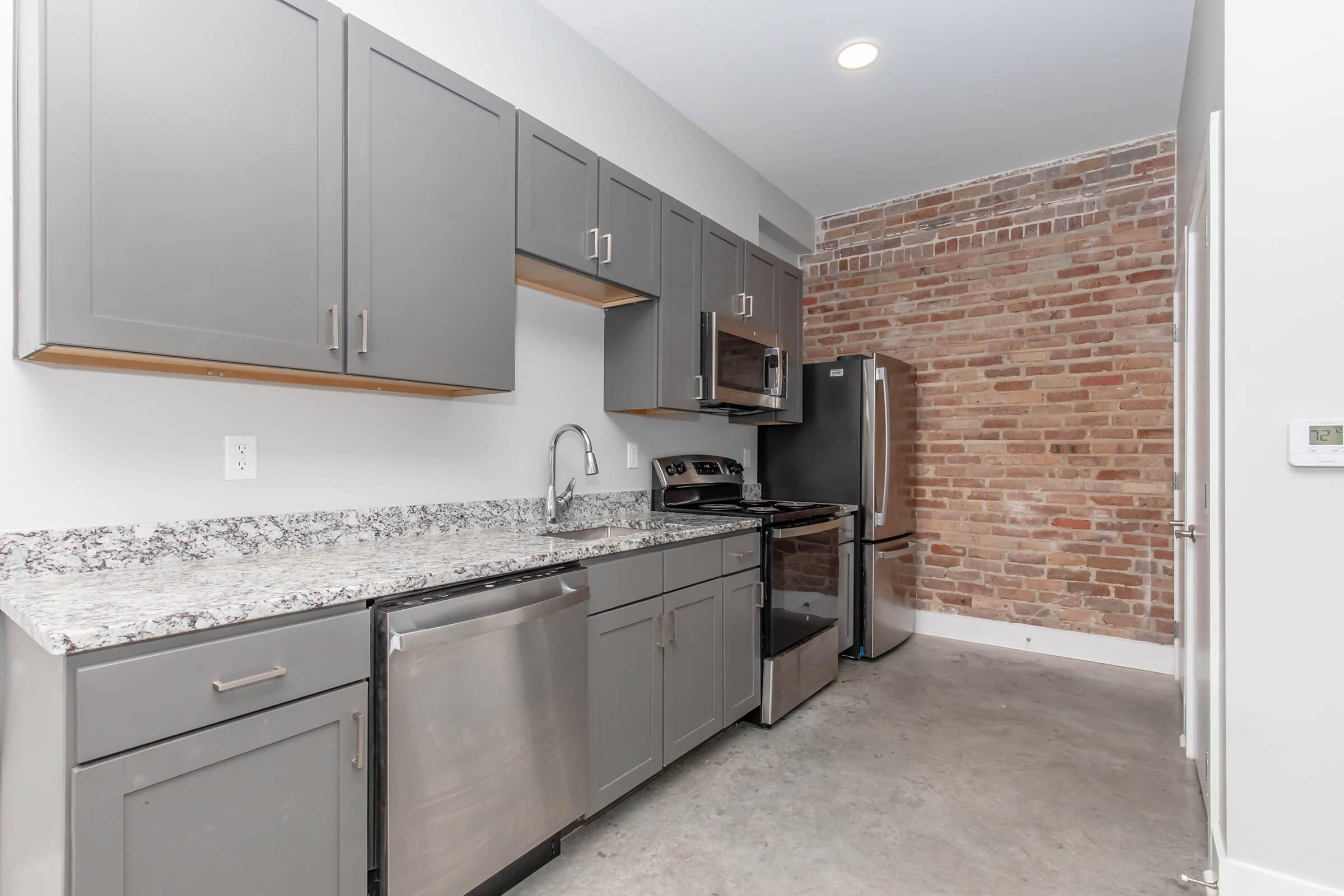 Modern kitchen featuring gray cabinetry, a granite countertop, stainless steel appliances including a refrigerator and stove, and a dishwasher. The walls are partially exposed brick, adding a rustic charm, with a concrete floor adding a contemporary touch. Brightly lit with overhead lighting.